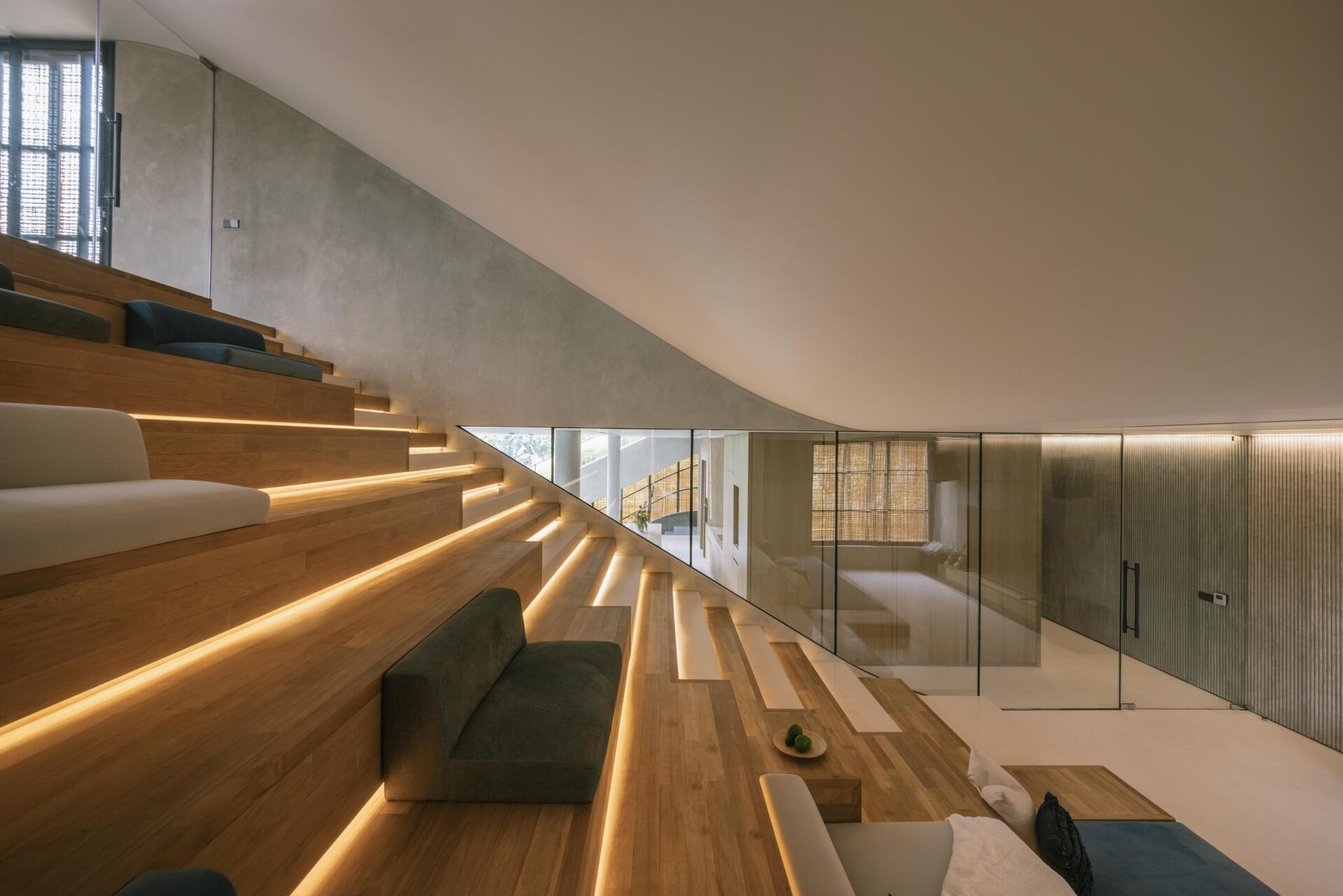 A relaxed entertainment discussion area features built-in stepped seating, a large screen, timber details and integrated lighting within Split Mass Residence in Indonesia.