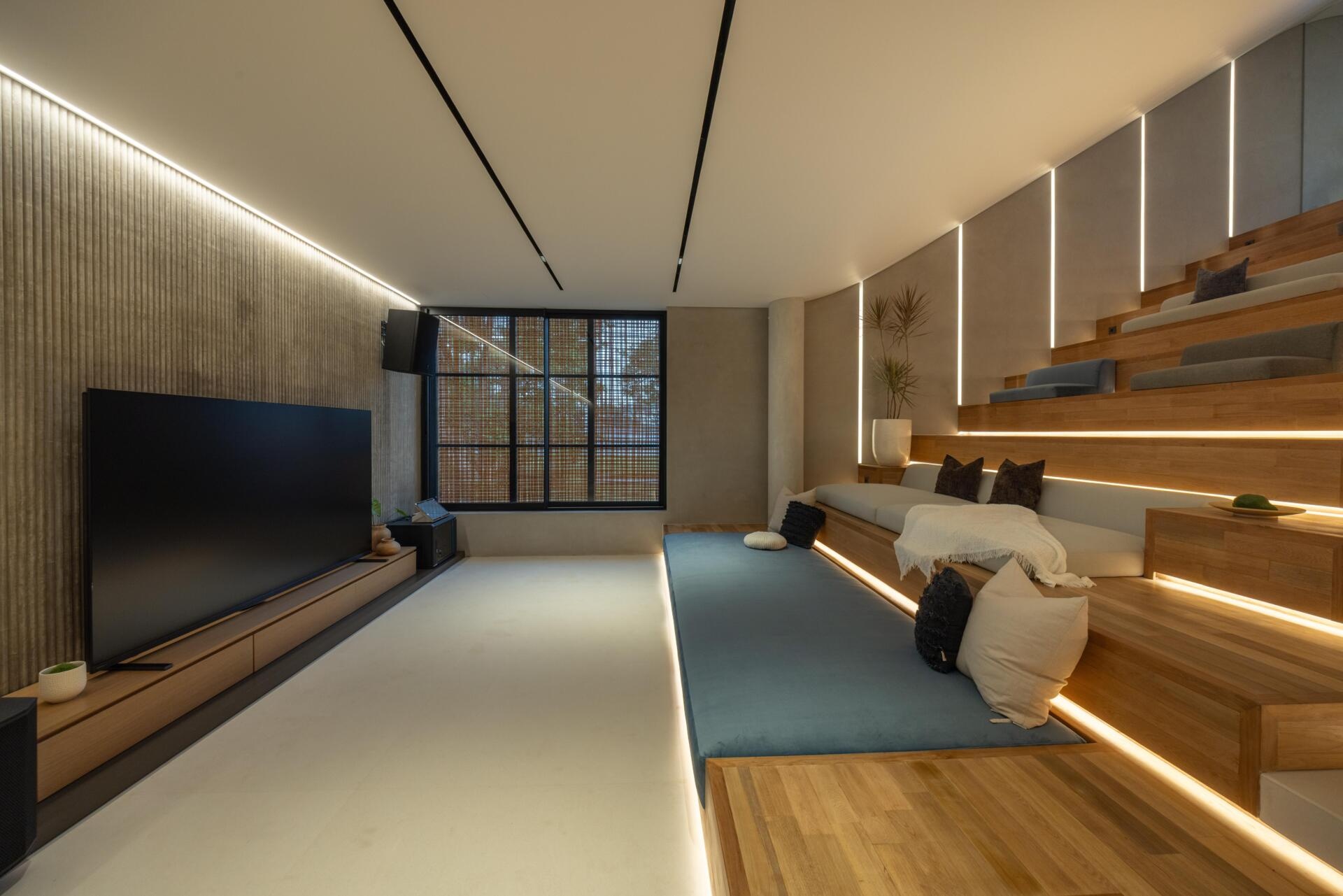 A relaxed entertainment discussion area features built-in stepped seating, a large screen, timber details and integrated lighting within Split Mass Residence in Indonesia.