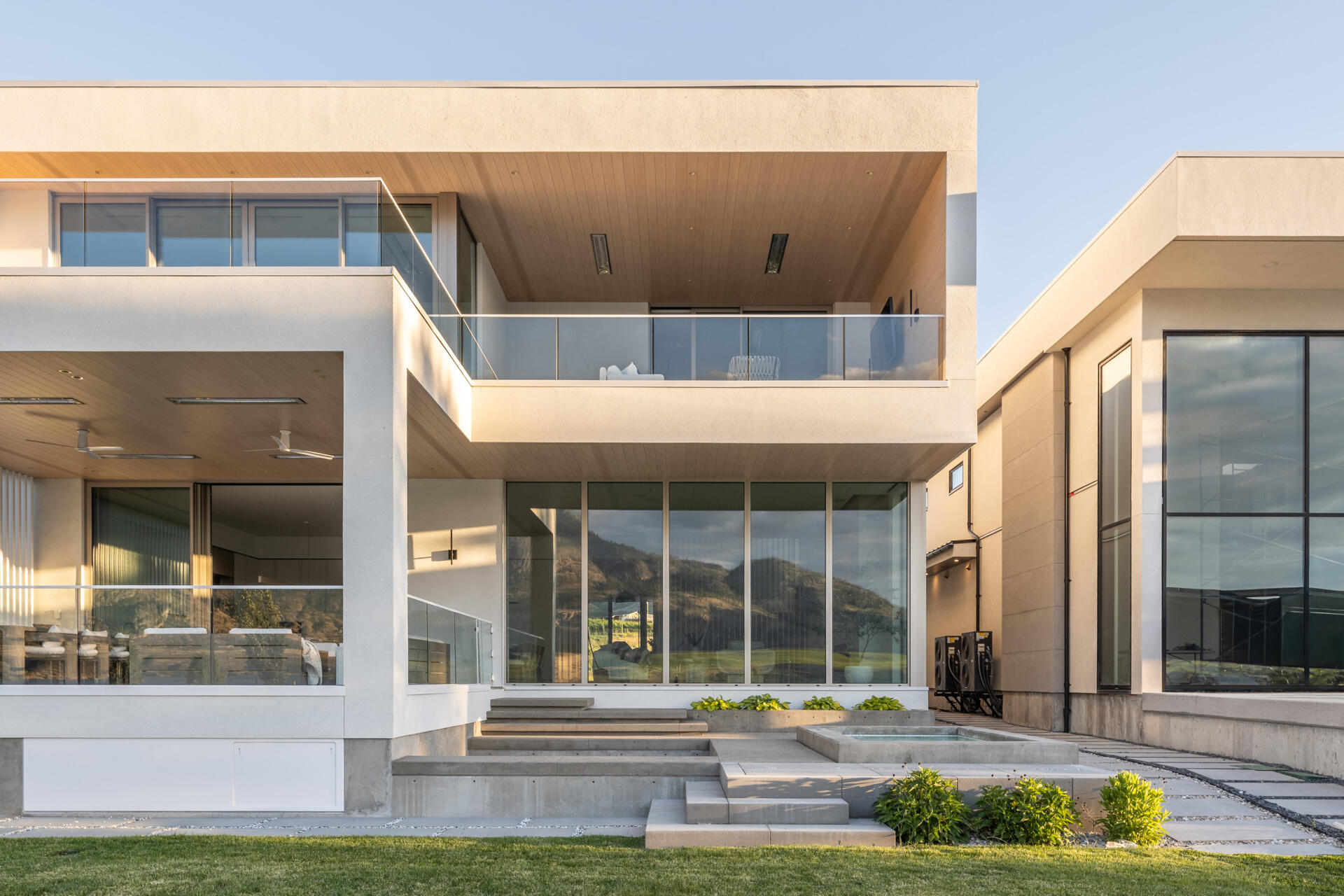 Explore the rear facade of this modern lake house, featuring white stucco, broad glass walls, warm wood ceilings, covered terraces, glass railings, and wide steps leading into the yard.