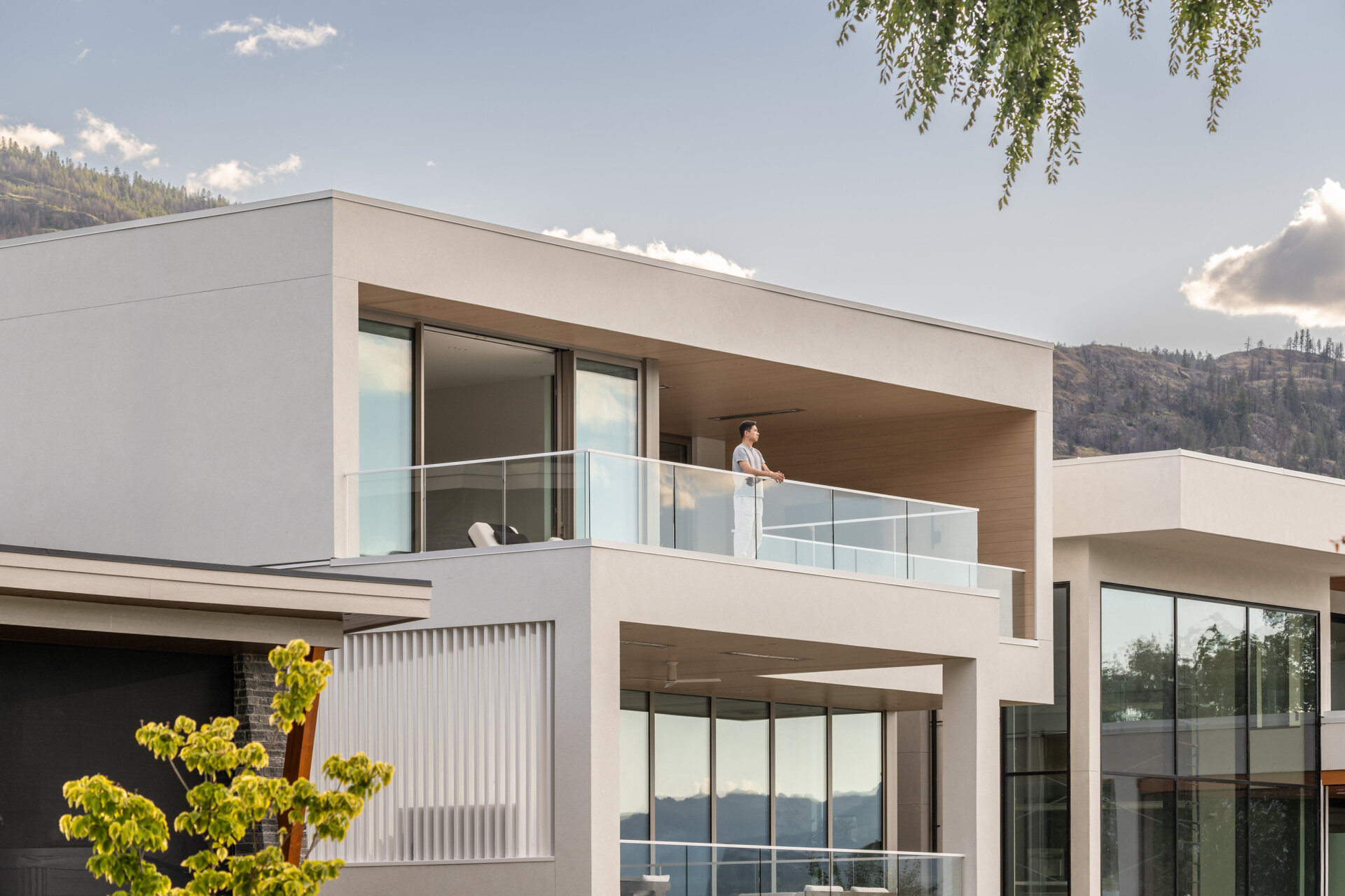 Explore the rear facade of this modern lake house, featuring white stucco, broad glass walls, warm wood ceilings, covered terraces, glass railings, and wide steps leading into the yard.