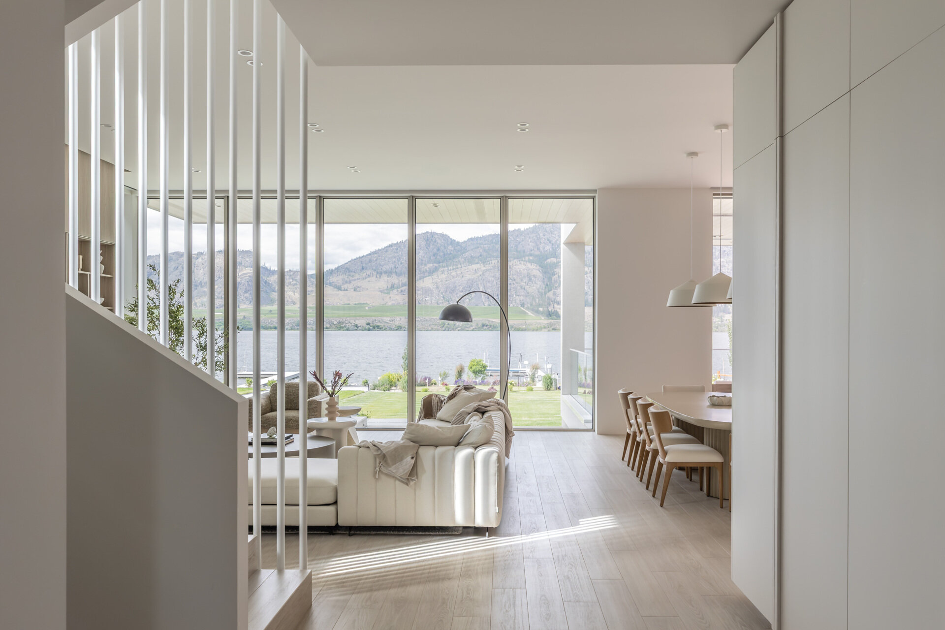 Take a look inside this modern lake house living room, where floor-to-ceiling glass frames lake and mountain views alongside pale wood paneling, neutral furniture, built-in shelving, and a long fireplace.