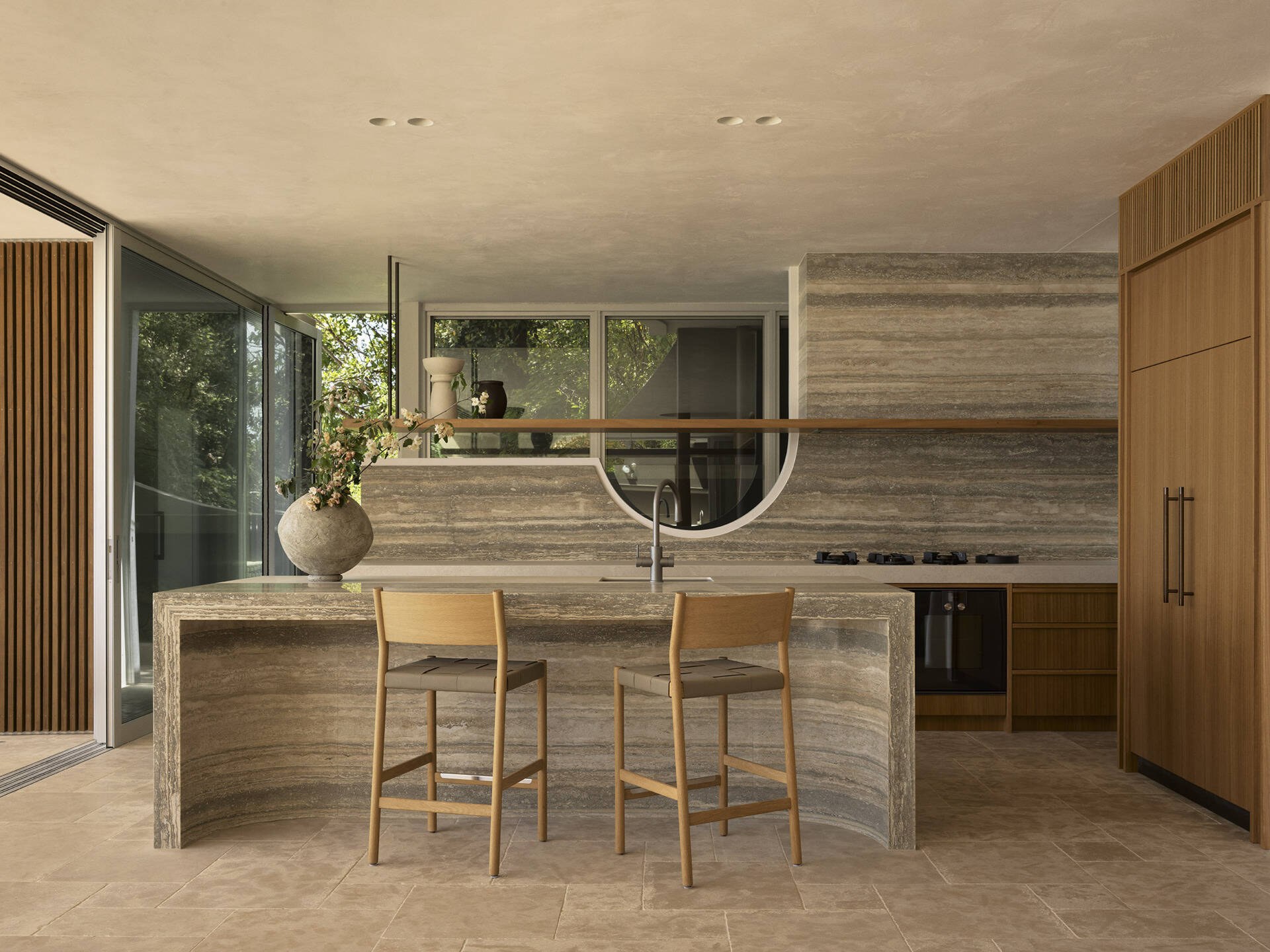 Step inside this Byron Bay kitchen featuring ocean-blue travertine, curved details, spliced stone and textured porcelain, paired with warm timber and expansive views across Wategos Beach.