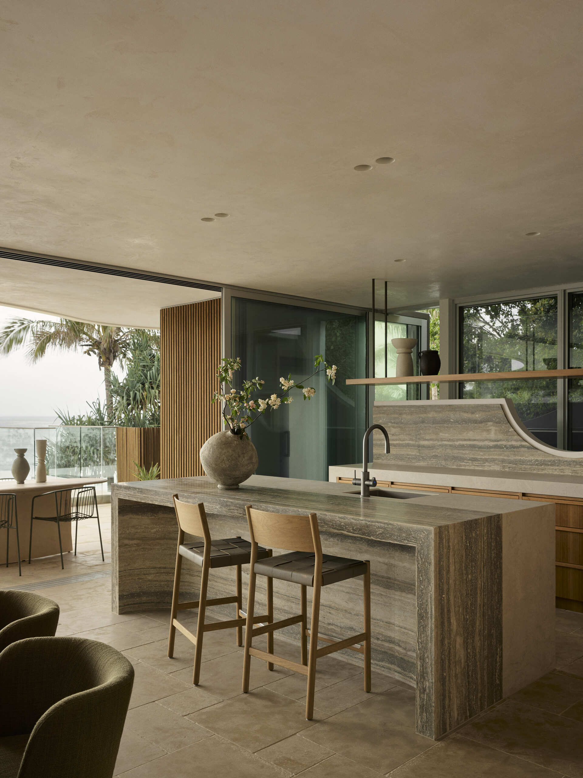 Step inside this Byron Bay kitchen featuring ocean-blue travertine, curved details, spliced stone and textured porcelain, paired with warm timber and expansive views across Wategos Beach.