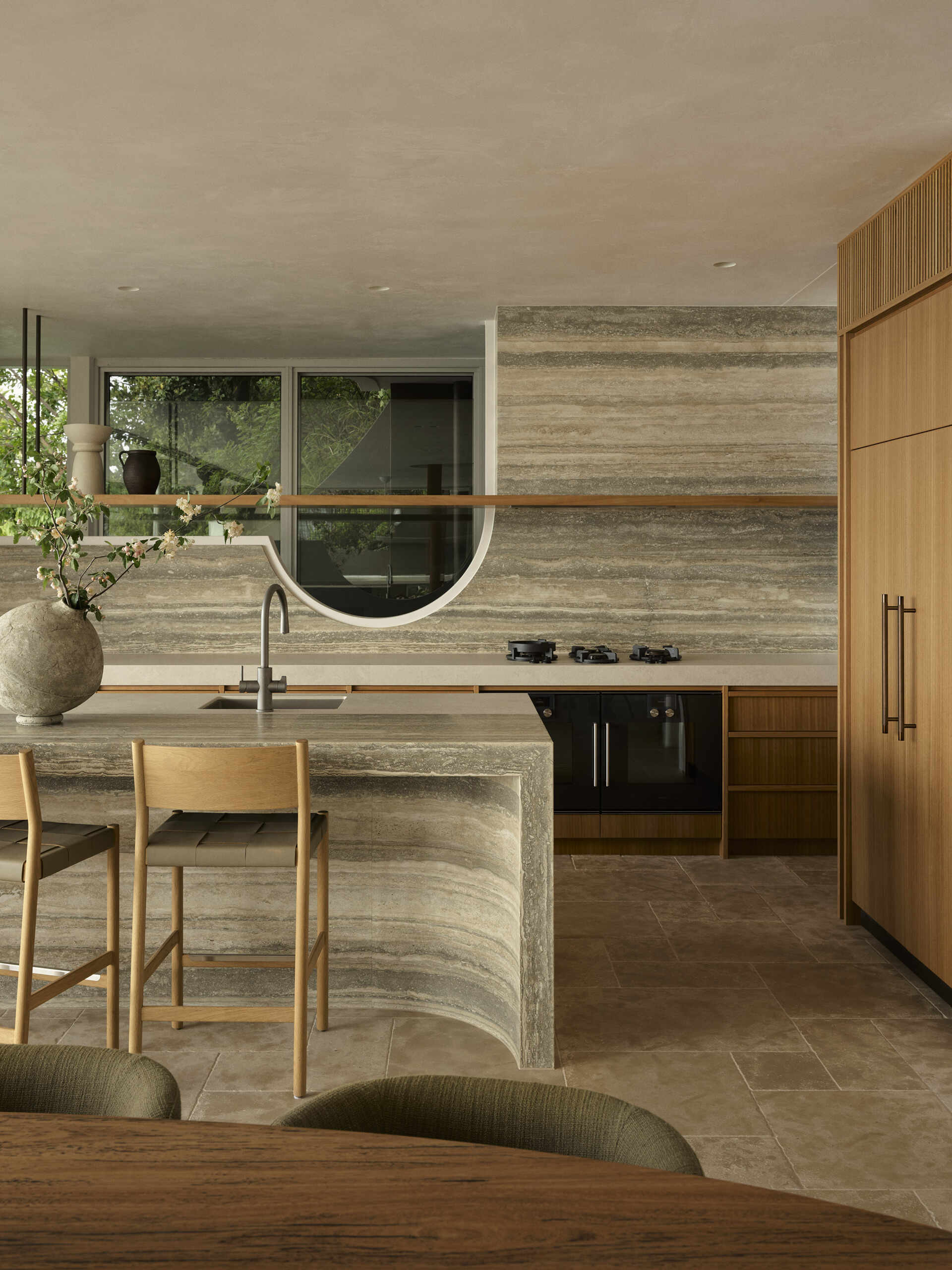 Step inside this Byron Bay kitchen featuring ocean-blue travertine, curved details, spliced stone and textured porcelain, paired with warm timber and expansive views across Wategos Beach.