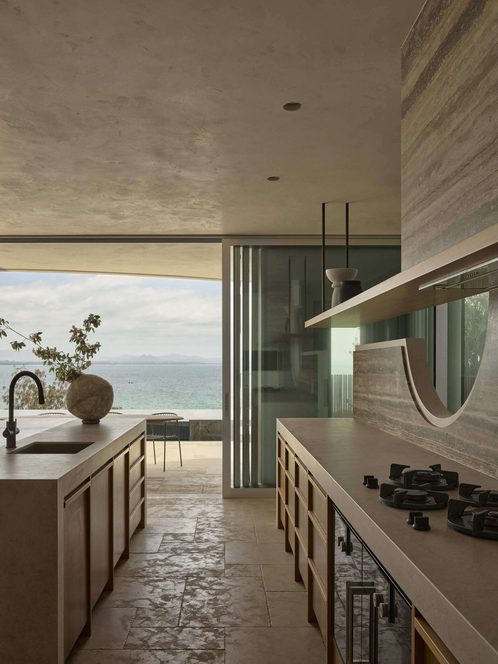 Step inside this Byron Bay kitchen featuring ocean-blue travertine, curved details, spliced stone and textured porcelain, paired with warm timber and expansive views across Wategos Beach.