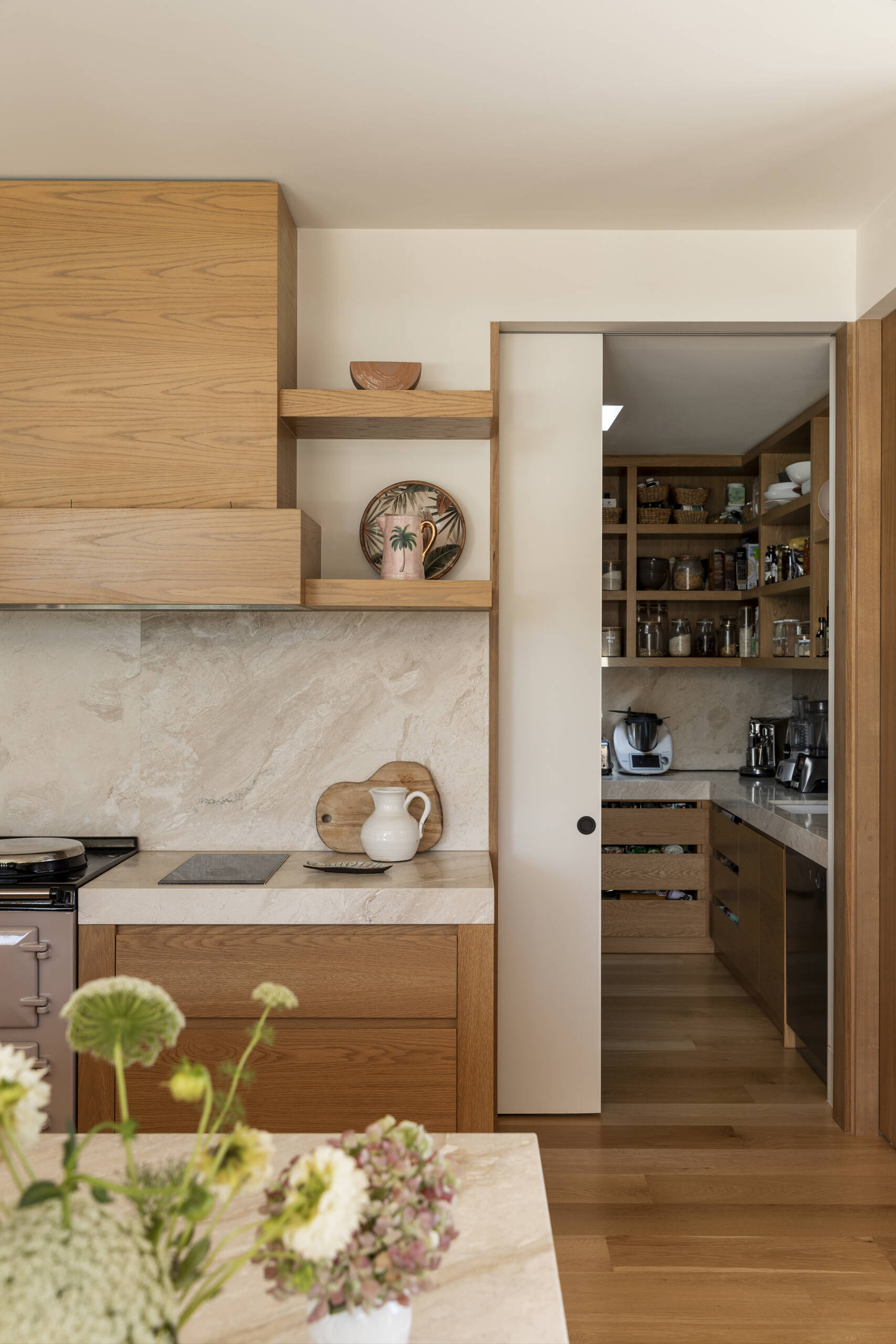 Natural wood cabinetry, woven pendant lights and softly veined stone give the kitchen and pantry a warm, relaxed character while keeping the look clean and contemporary. The oversized island provides plenty of space for cooking, casual meals and gathering with friends, with woven counter stools adding comfort and texture.