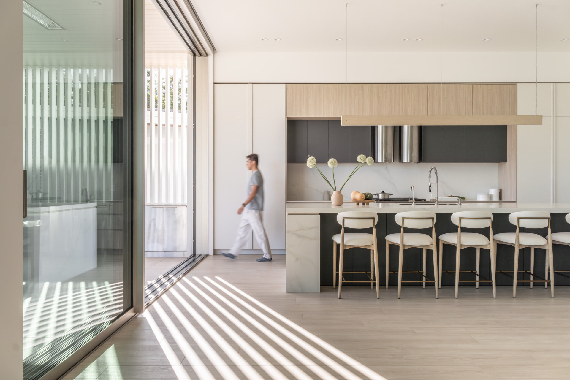 Explore this light-filled modern kitchen featuring pale wood cabinetry, crisp white surfaces, a large island with seating for five, and a wide opening that brings the outdoors inside.