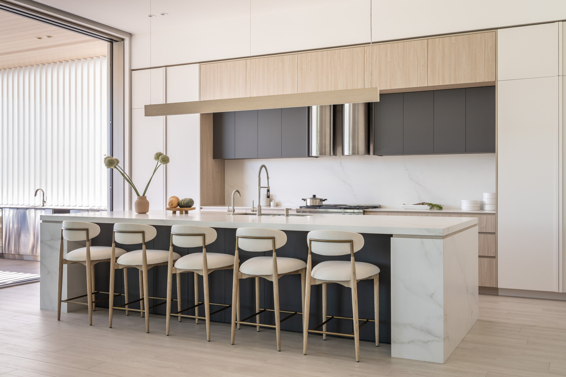 Explore this light-filled modern kitchen featuring pale wood cabinetry, crisp white surfaces, a large island with seating for five, and a wide opening that brings the outdoors inside.