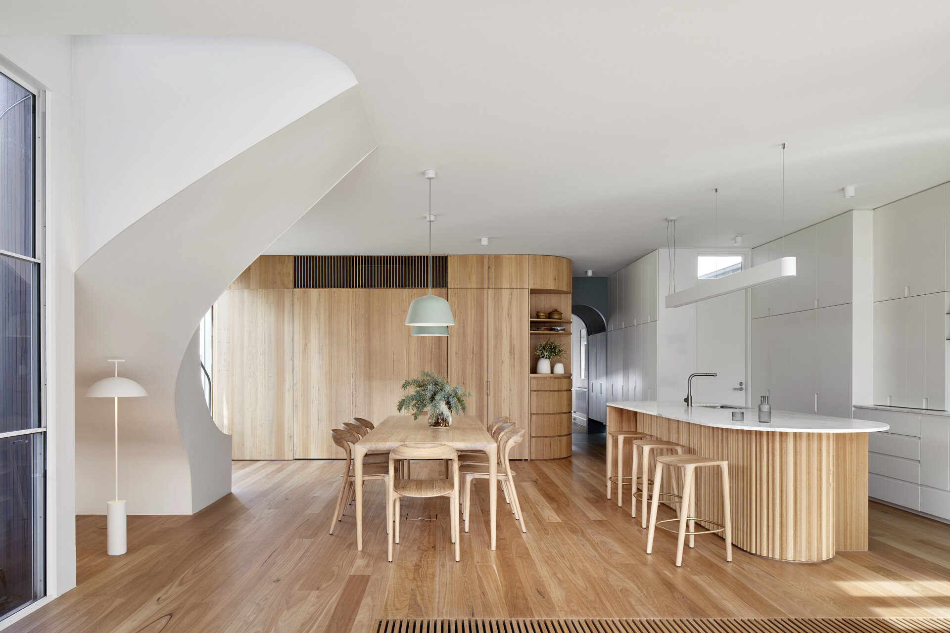 The dining area flows directly from the kitchen, with warm Blackbutt floors, white walls, and curved furniture.