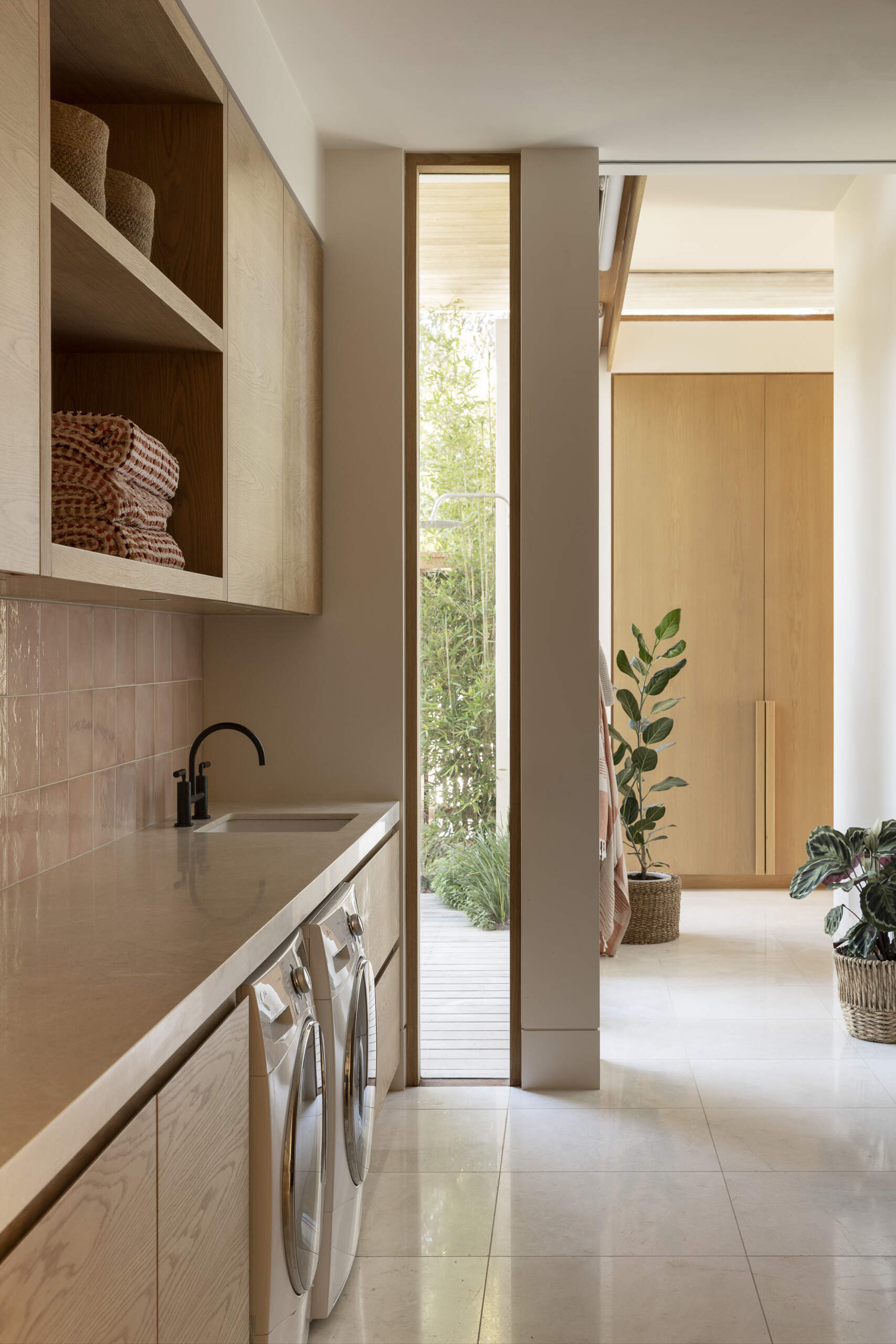 The laundry combines soft wood cabinetry with warm stone counters and glossy blush-toned tile, giving this practical room a calm, refined character. Open shelving keeps baskets and folded linens close at hand, while the long counter provides plenty of space for sorting and folding. A slim floor-to-ceiling window brings natural light into the room along with a glimpse of greenery outside.
