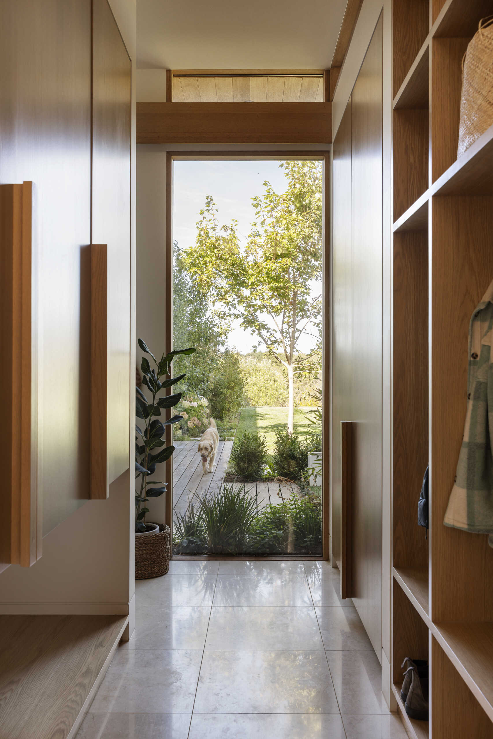 The mud room pairs streamlined wood cabinetry with open cubbies and oversized handles. A pale stone floor keeps the palette light, while woven baskets and a leafy potted plant add texture. A full-height glass door frames the garden beyond, bringing daylight into the narrow entrance and creating a visual link with the greenery outside.