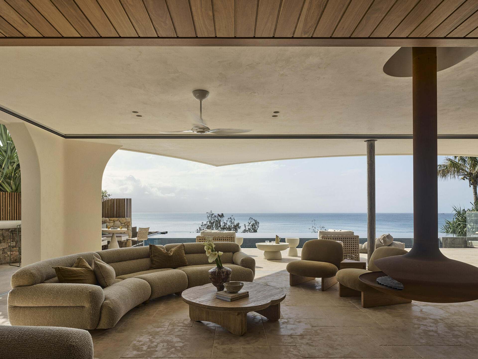 Warm stone floors, creamy textured walls, rich timber and curved furniture create a relaxed living and dining space overlooking the ocean, with wide openings extending the room onto the terrace.