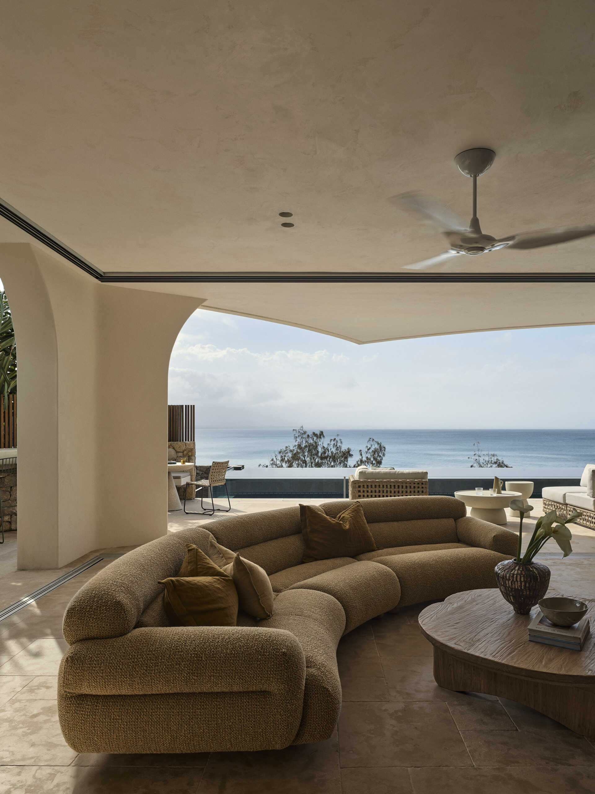 Warm stone floors, creamy textured walls, rich timber and curved furniture create a relaxed living and dining space overlooking the ocean, with wide openings extending the room onto the terrace.