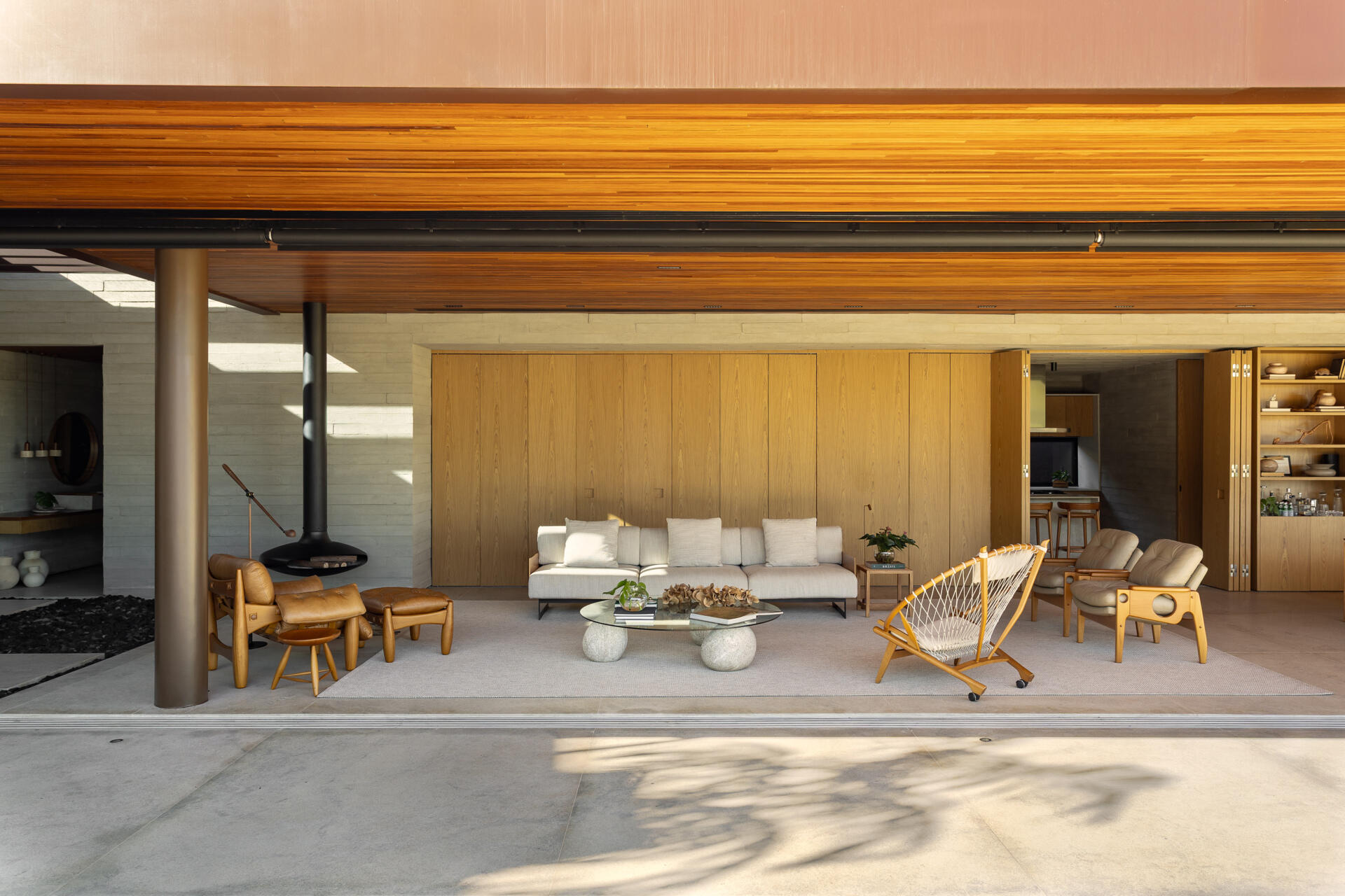 Explore the living room of Casa da Mata, featuring warm wood, pale concrete, cognac leather seating, a sculptural fireplace, textured furnishings and wide openings overlooking lush tropical planting.