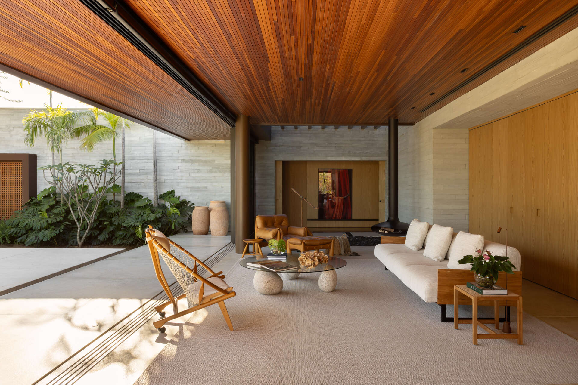 Explore the living room of Casa da Mata, featuring warm wood, pale concrete, cognac leather seating, a sculptural fireplace, textured furnishings and wide openings overlooking lush tropical planting.