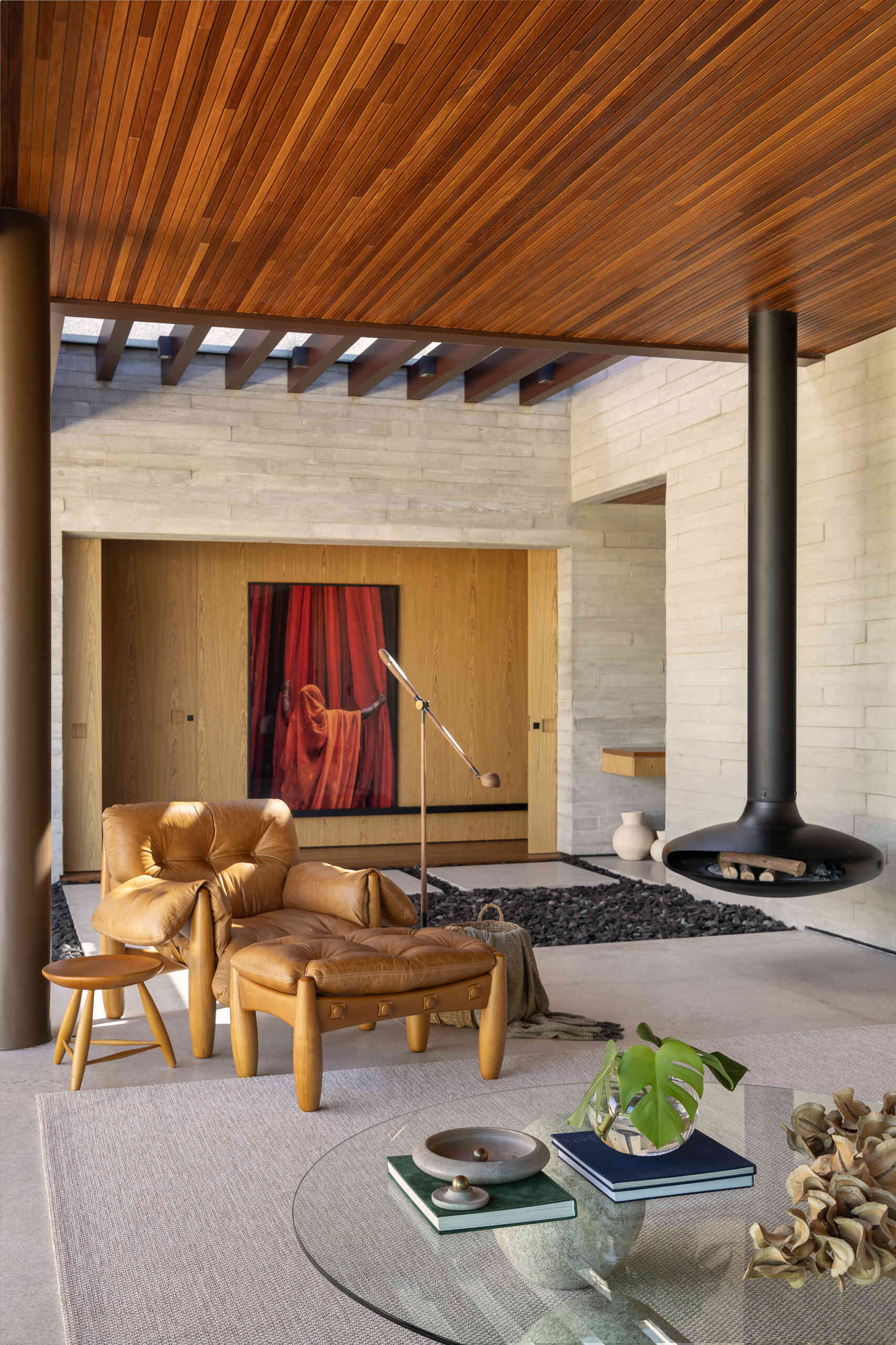 Explore the living room of Casa da Mata, featuring warm wood, pale concrete, cognac leather seating, a sculptural fireplace, textured furnishings and wide openings overlooking lush tropical planting.