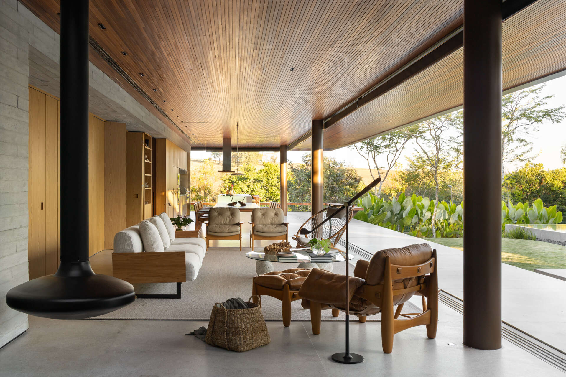 Explore the living room of Casa da Mata, featuring warm wood, pale concrete, cognac leather seating, a sculptural fireplace, textured furnishings and wide openings overlooking lush tropical planting.