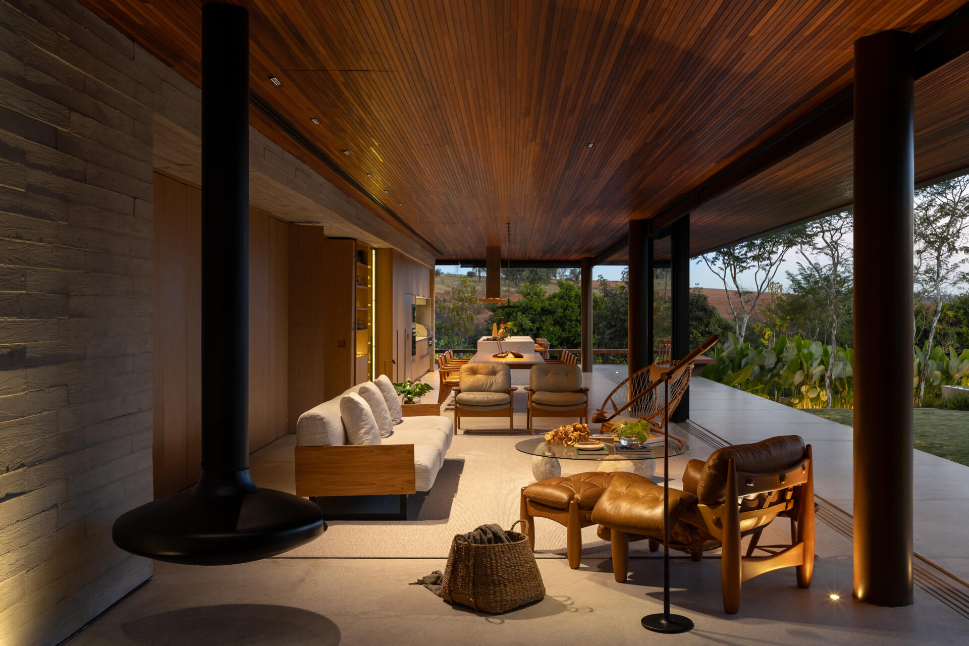 Explore the living room of Casa da Mata, featuring warm wood, pale concrete, cognac leather seating, a sculptural fireplace, textured furnishings and wide openings overlooking lush tropical planting.