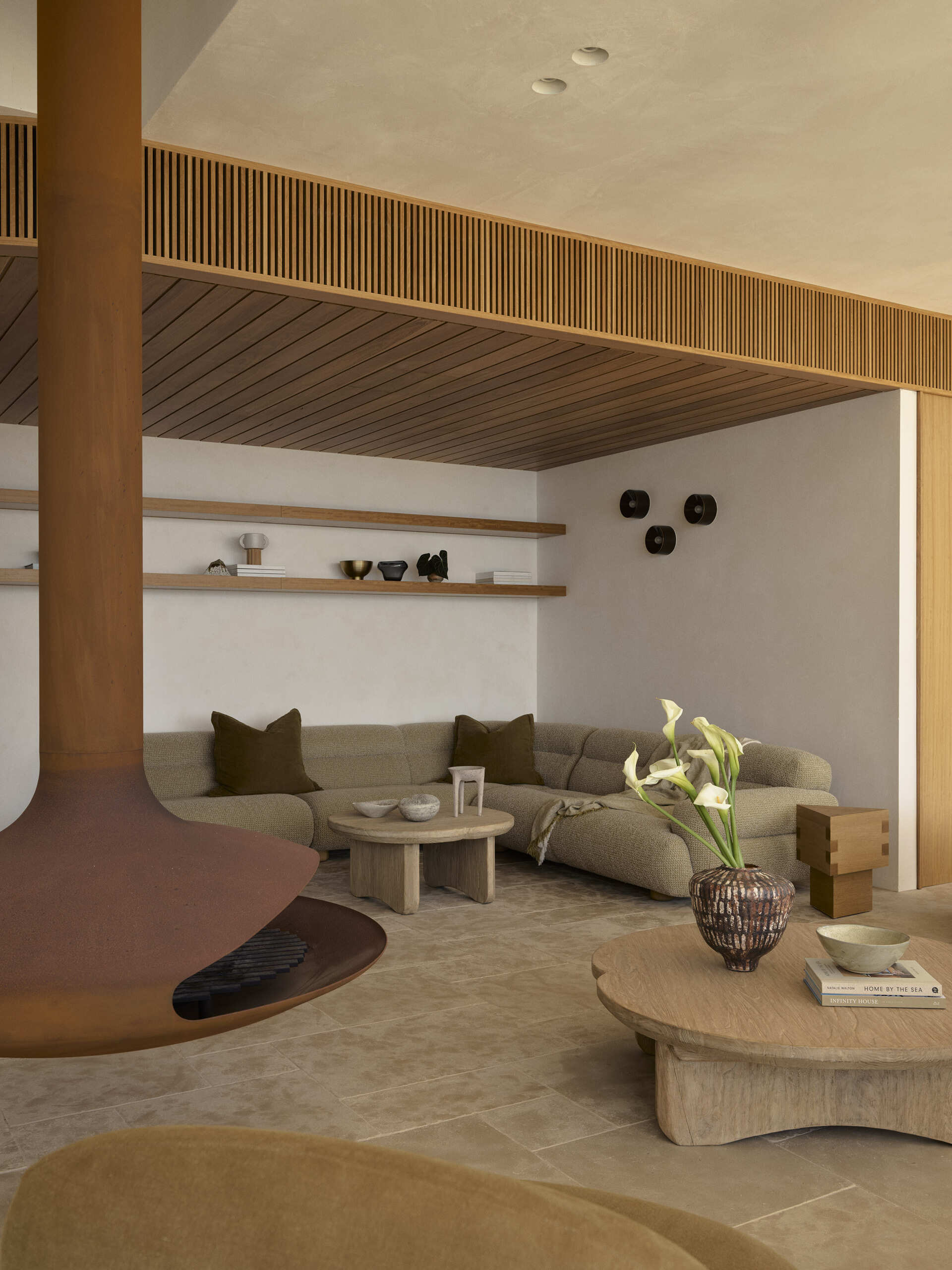 Warm stone floors, creamy textured walls, rich timber and curved furniture create a relaxed living and dining space overlooking the ocean, with wide openings extending the room onto the terrace.