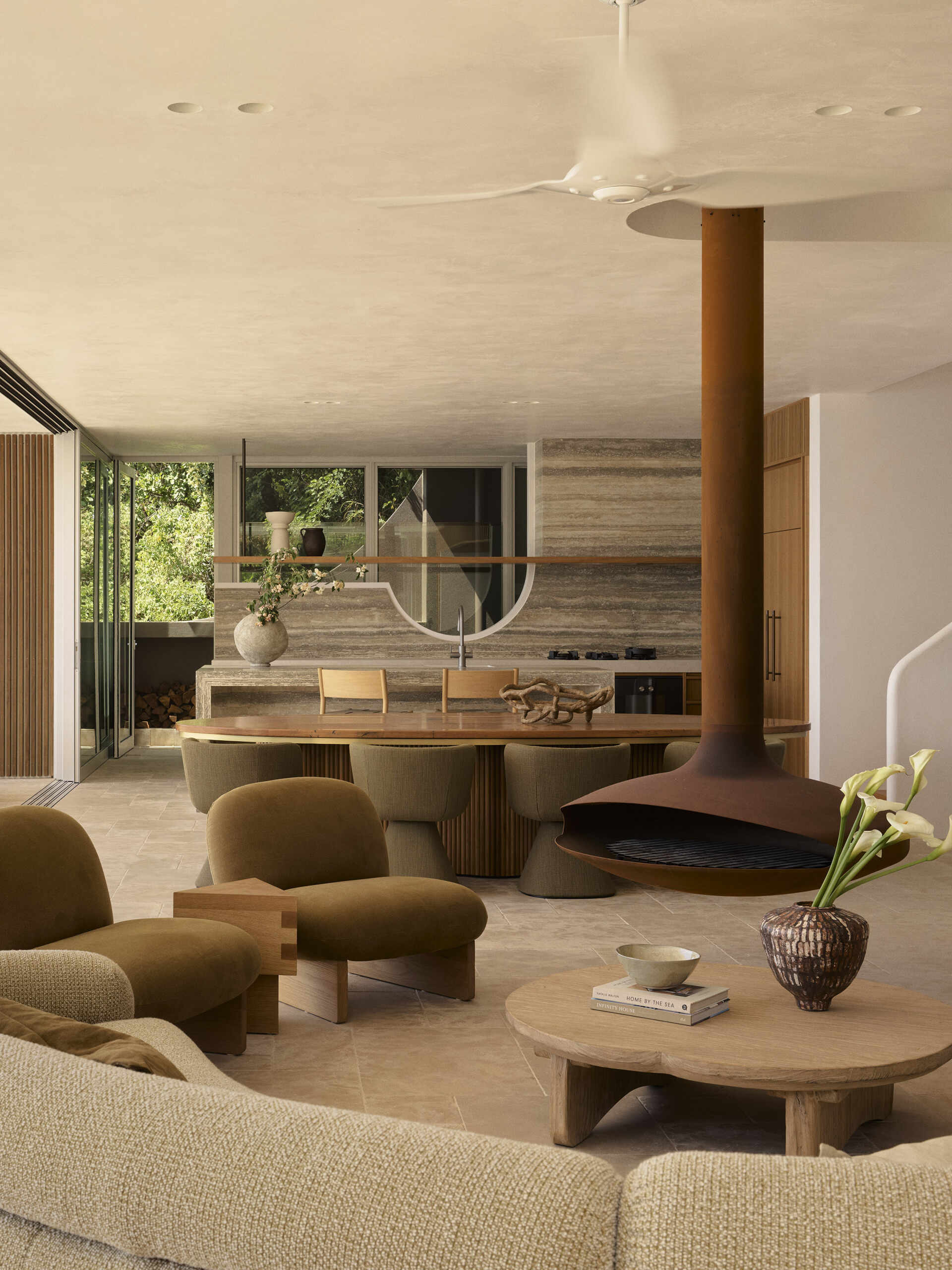 Warm stone floors, creamy textured walls, rich timber and curved furniture create a relaxed living and dining space overlooking the ocean, with wide openings extending the room onto the terrace.