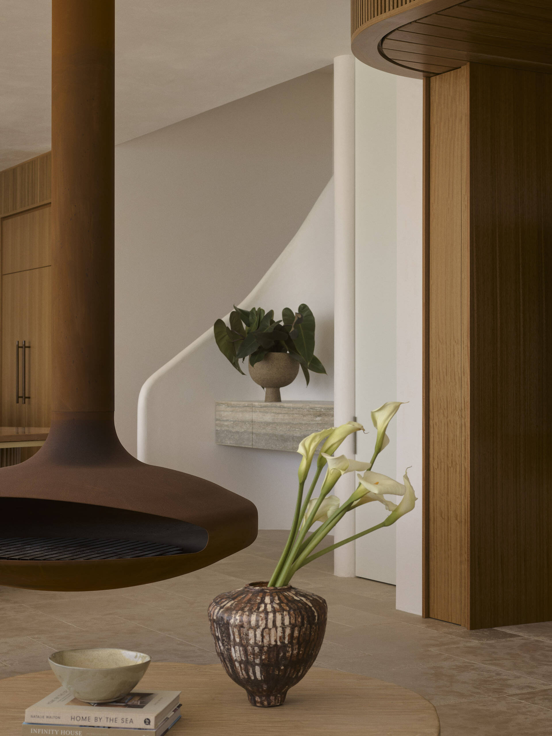 Warm stone floors, creamy textured walls, rich timber and curved furniture create a relaxed living and dining space overlooking the ocean, with wide openings extending the room onto the terrace.