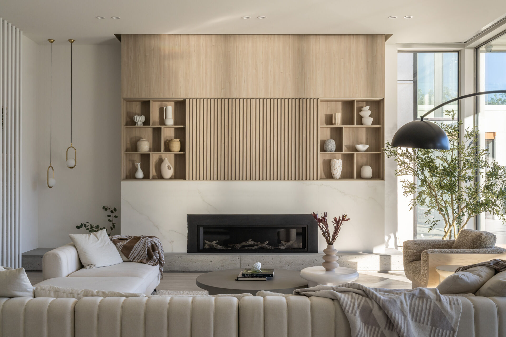 Take a look inside this modern lake house living room, where floor-to-ceiling glass frames lake and mountain views alongside pale wood paneling, neutral furniture, built-in shelving, and a long fireplace.