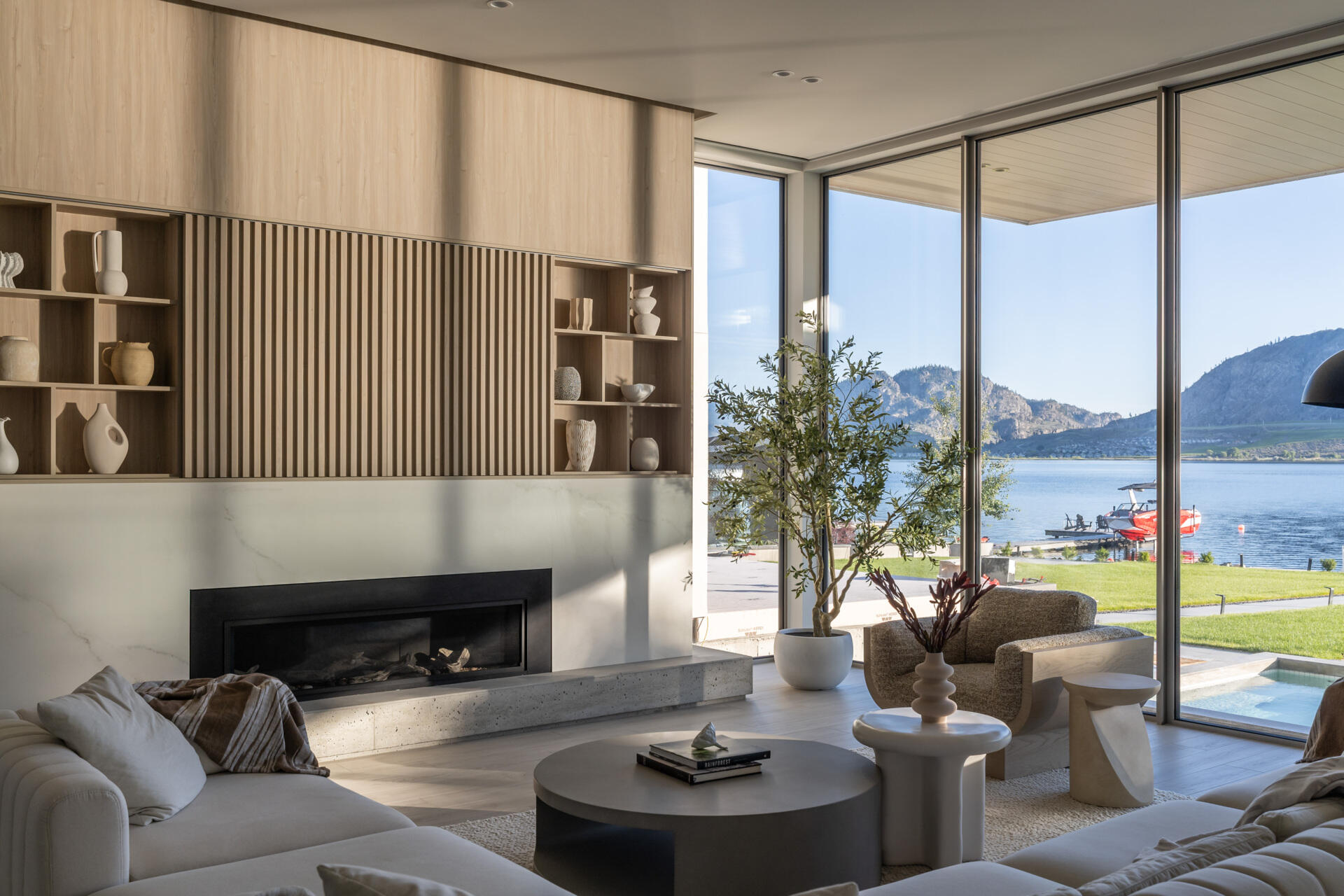 Take a look inside this modern lake house living room, where floor-to-ceiling glass frames lake and mountain views alongside pale wood paneling, neutral furniture, built-in shelving, and a long fireplace.