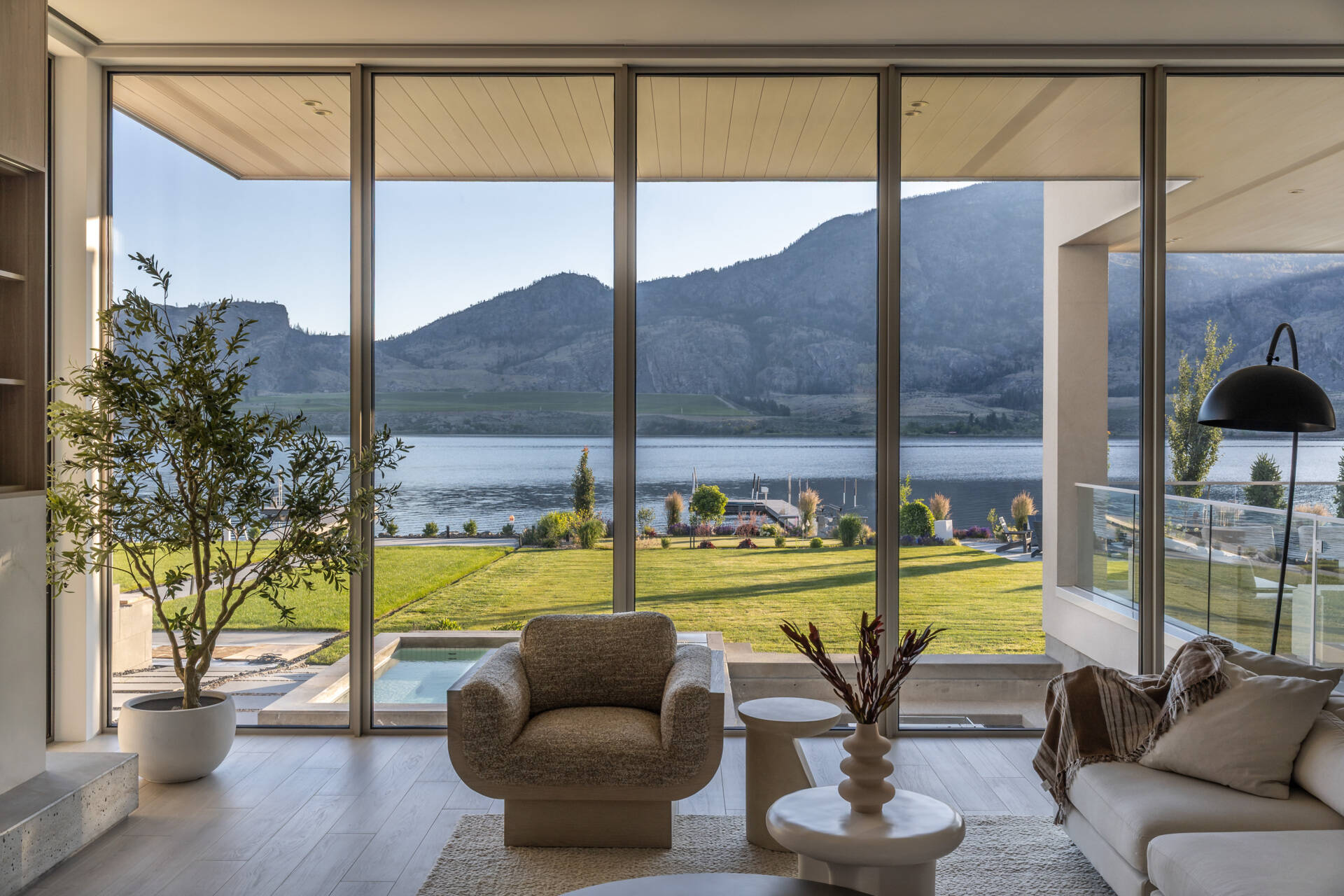 Take a look inside this modern lake house living room, where floor-to-ceiling glass frames lake and mountain views alongside pale wood paneling, neutral furniture, built-in shelving, and a long fireplace.