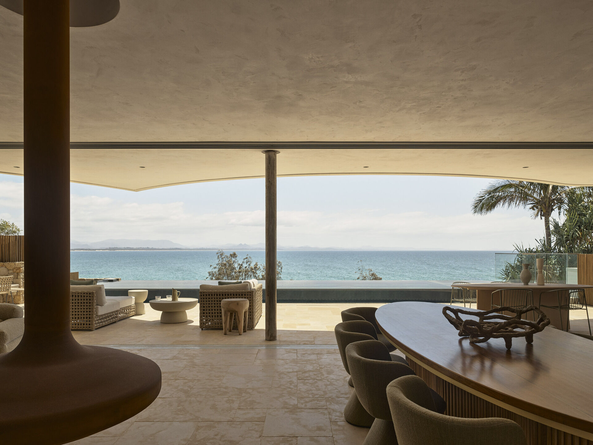 Warm stone floors, creamy textured walls, rich timber and curved furniture create a relaxed living and dining space overlooking the ocean, with wide openings extending the room onto the terrace.