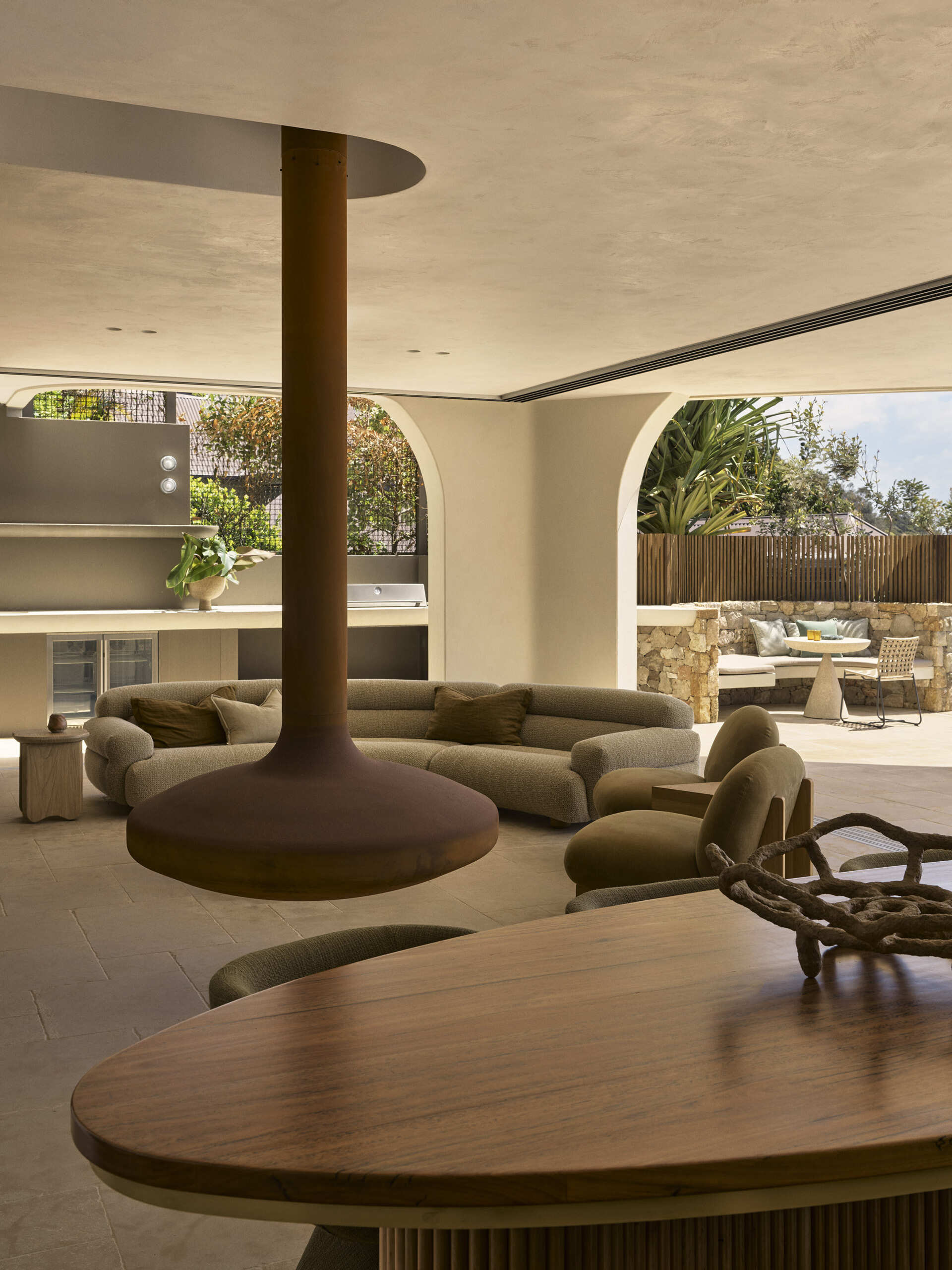 Warm stone floors, creamy textured walls, rich timber and curved furniture create a relaxed living and dining space overlooking the ocean, with wide openings extending the room onto the terrace.