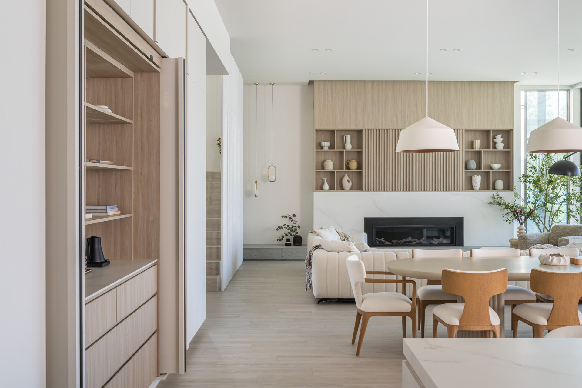 See how this modern kitchen and dining space combines pale wood finishes, white cabinetry, sculptural lighting, and a clever hidden storage wall that conceals shelving, drawers, and appliances.