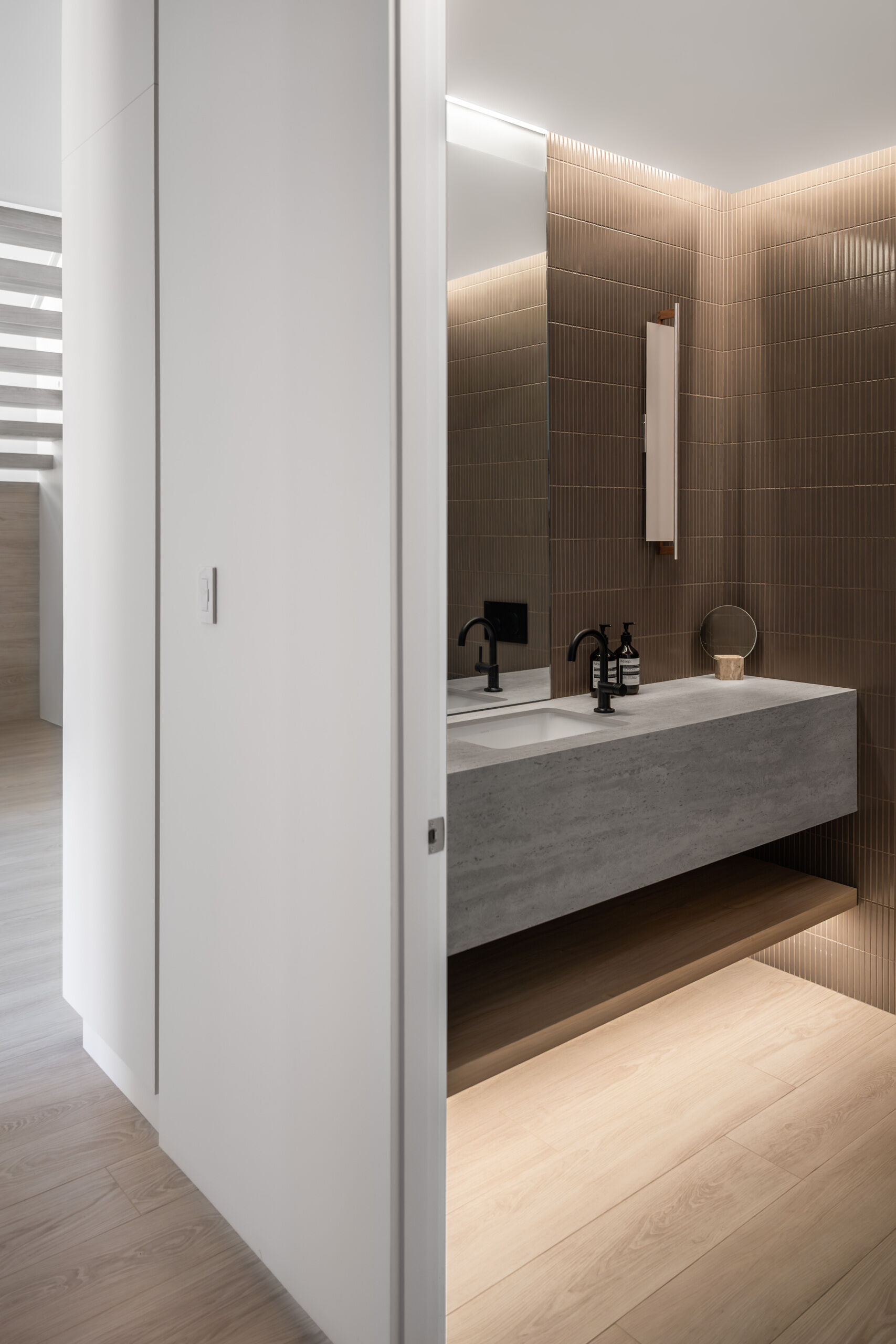 Get inspiration from this modern powder room featuring deep brown vertical tile, a floating gray stone vanity, pale wood flooring, concealed lighting, and streamlined black and wood details.
