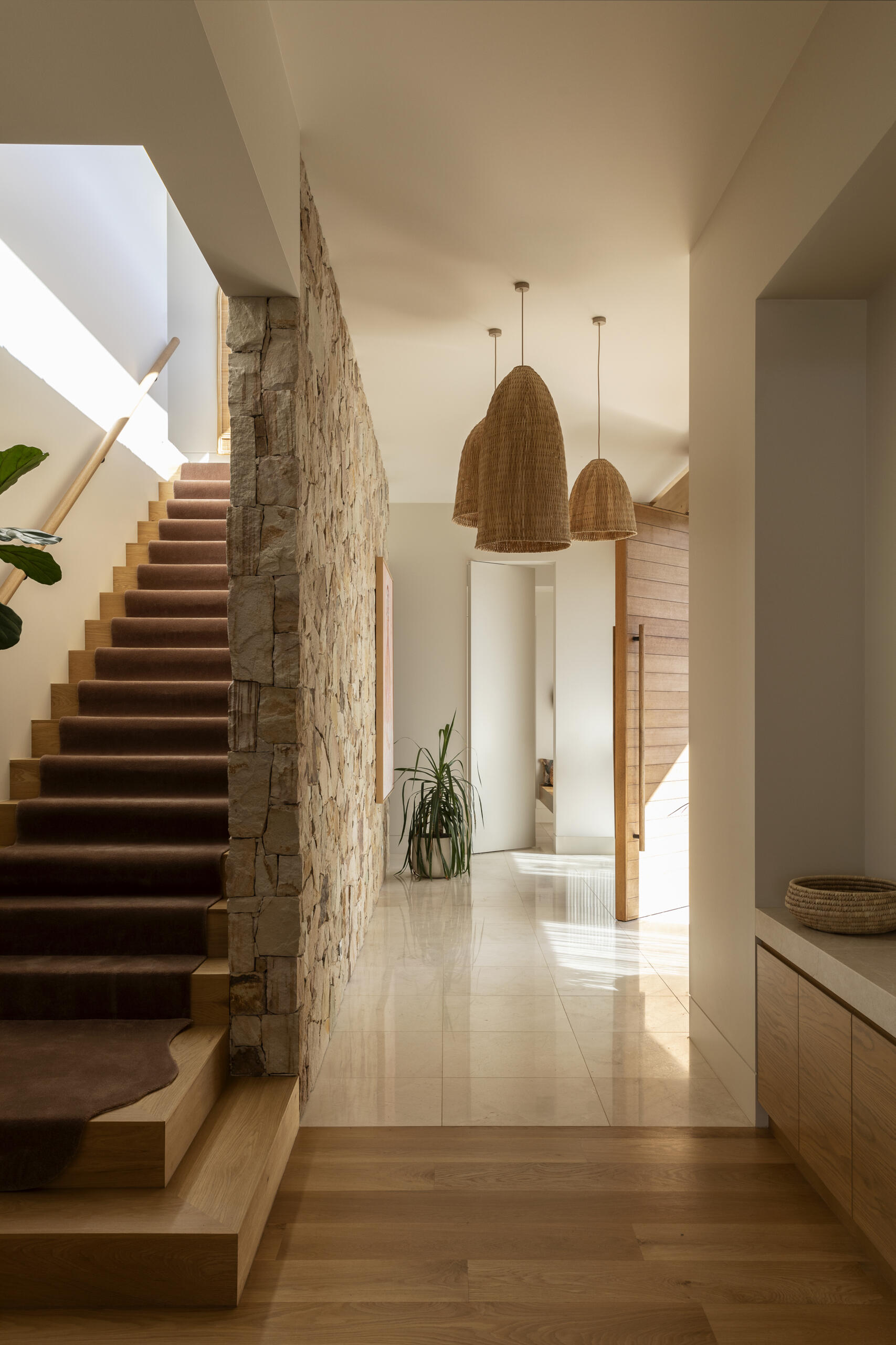 A rugged stone wall partially hides the staircase from the entryway. Sunlight from above creates shifting patterns across the pale stone floor, adding movement to the natural material palette.
