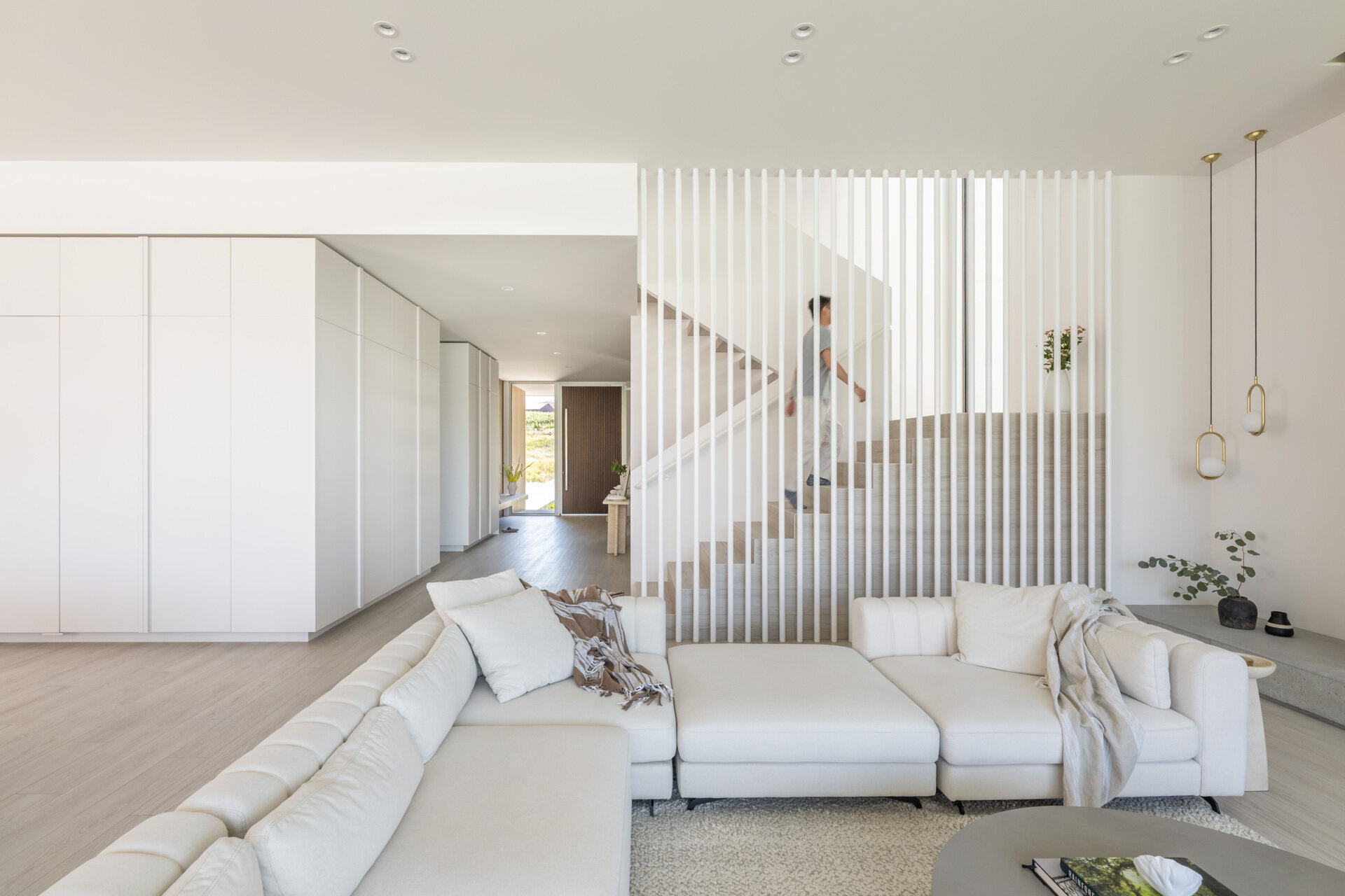 See how this modern staircase becomes part of an open living space, combining pale wood floors, slim vertical slats, built-in storage, delicate pendant lights, and a soft neutral palette.