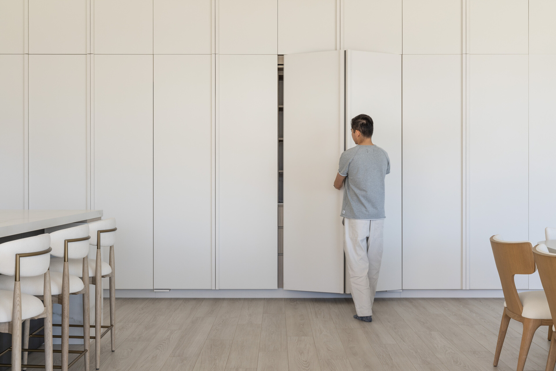 See how this modern kitchen and dining space combines pale wood finishes, white cabinetry, sculptural lighting, and a clever hidden storage wall that conceals shelving, drawers, and appliances.