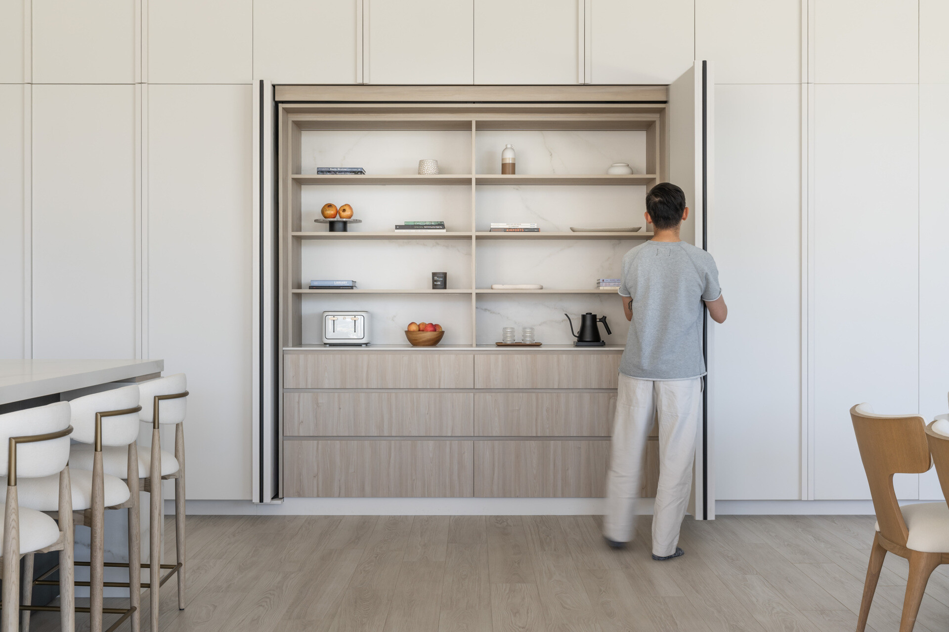 See how this modern kitchen and dining space combines pale wood finishes, white cabinetry, sculptural lighting, and a clever hidden storage wall that conceals shelving, drawers, and appliances.