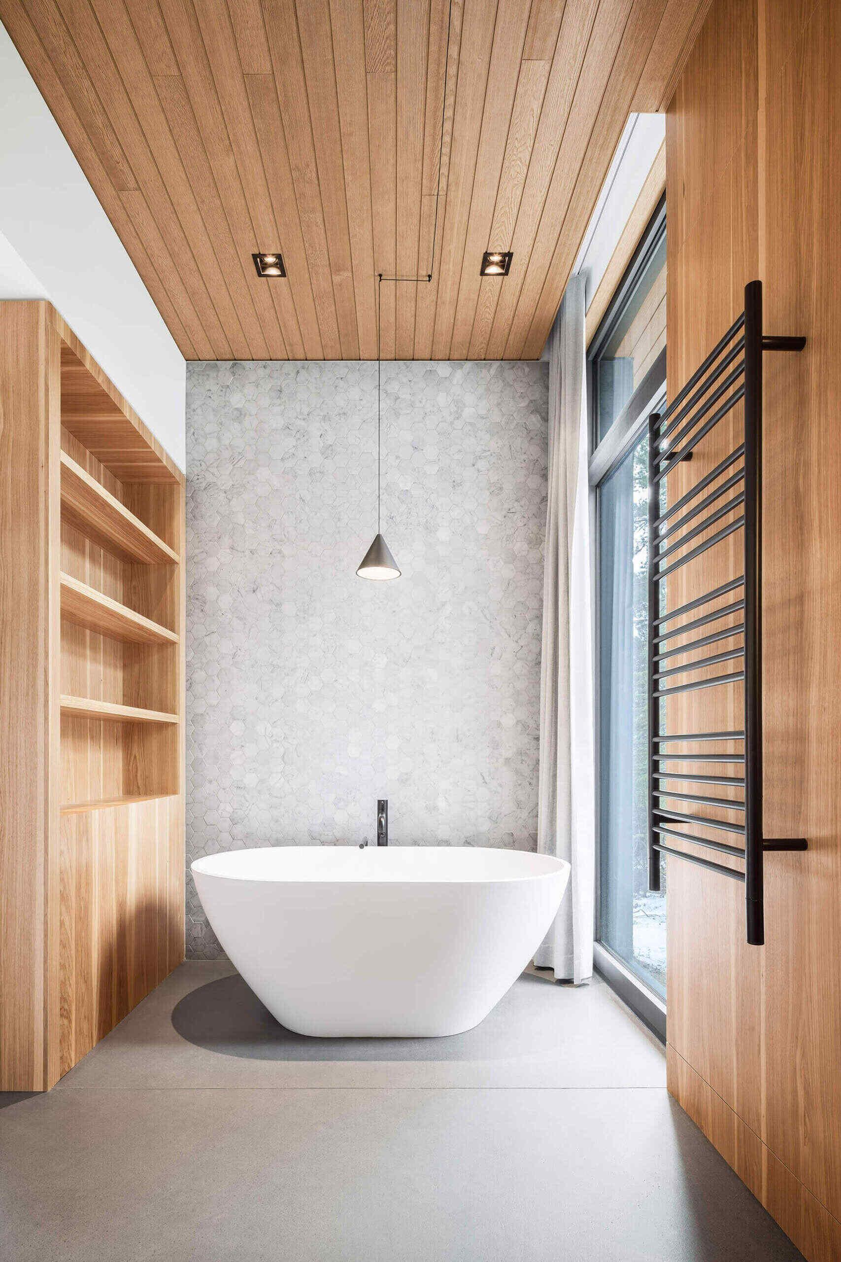 A white freestanding bathtub, grey tile, open shelving and a curtained window create a simple contemporary bathroom inside Long Horizontals in Québec.