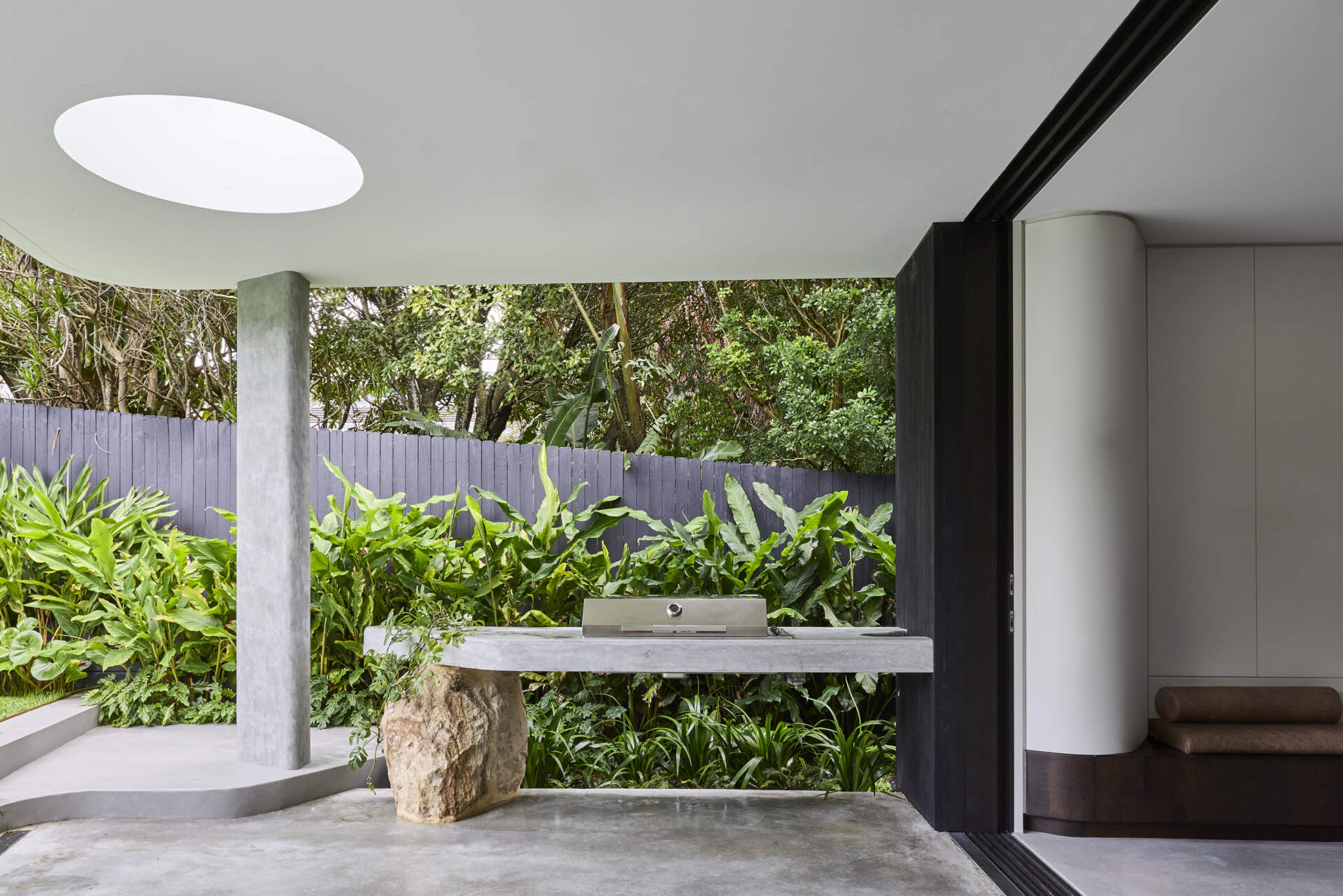 Discover the outdoor kitchen at Sasso Nero, with a long concrete counter, integrated barbecue and natural stone support beneath a covered section of the house. Lush garden planting surrounds the space.