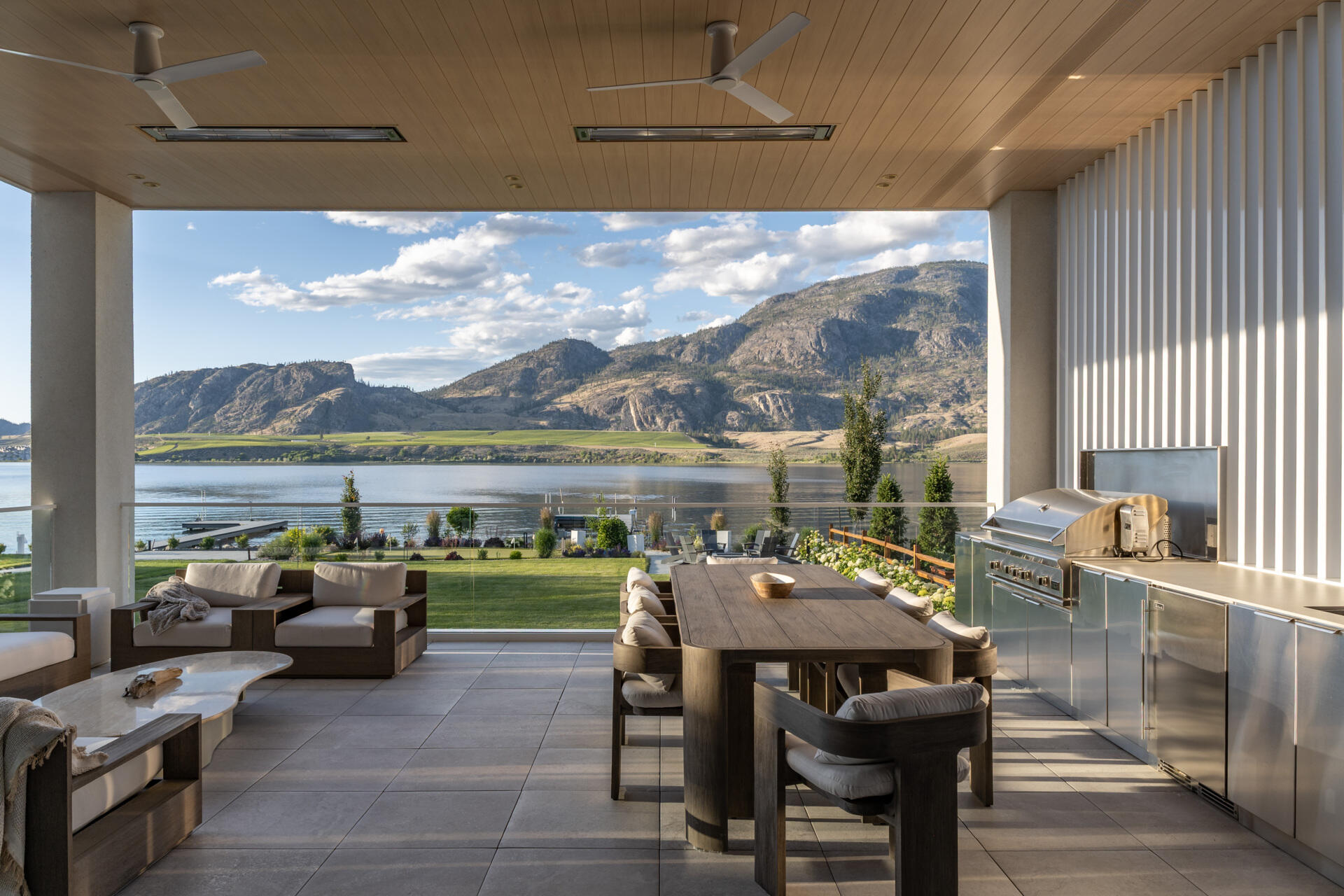 Discover this covered outdoor living area with sweeping lake and mountain views, a long dining table, comfortable lounge seating, outdoor kitchen, ceiling fans, heaters, and slatted screening.