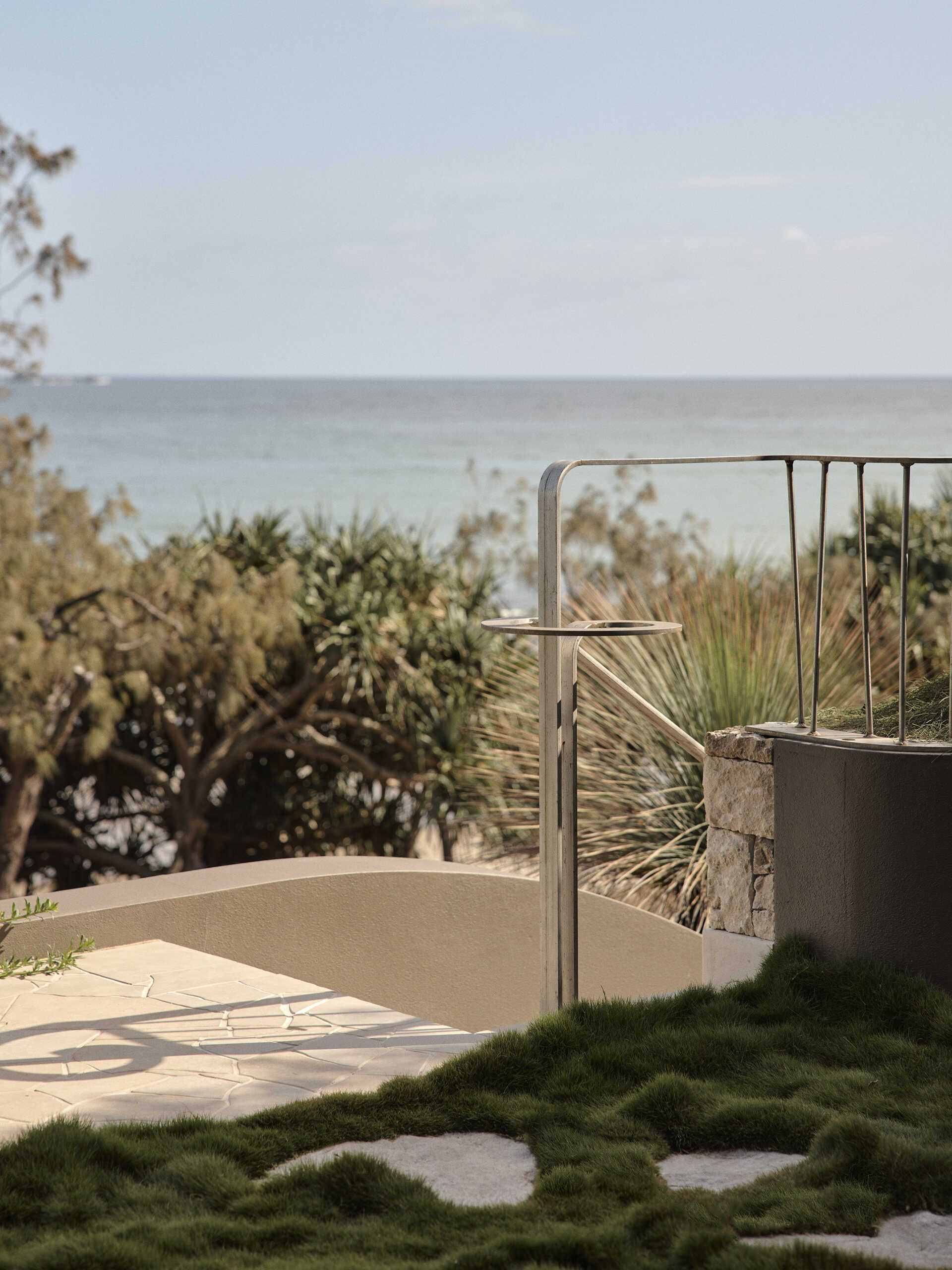 Curved taupe stucco walls, pale stone steps and lush planting create a sculptural entrance to this Byron Bay home, softened by a mature grass tree and touches of purple flowers.