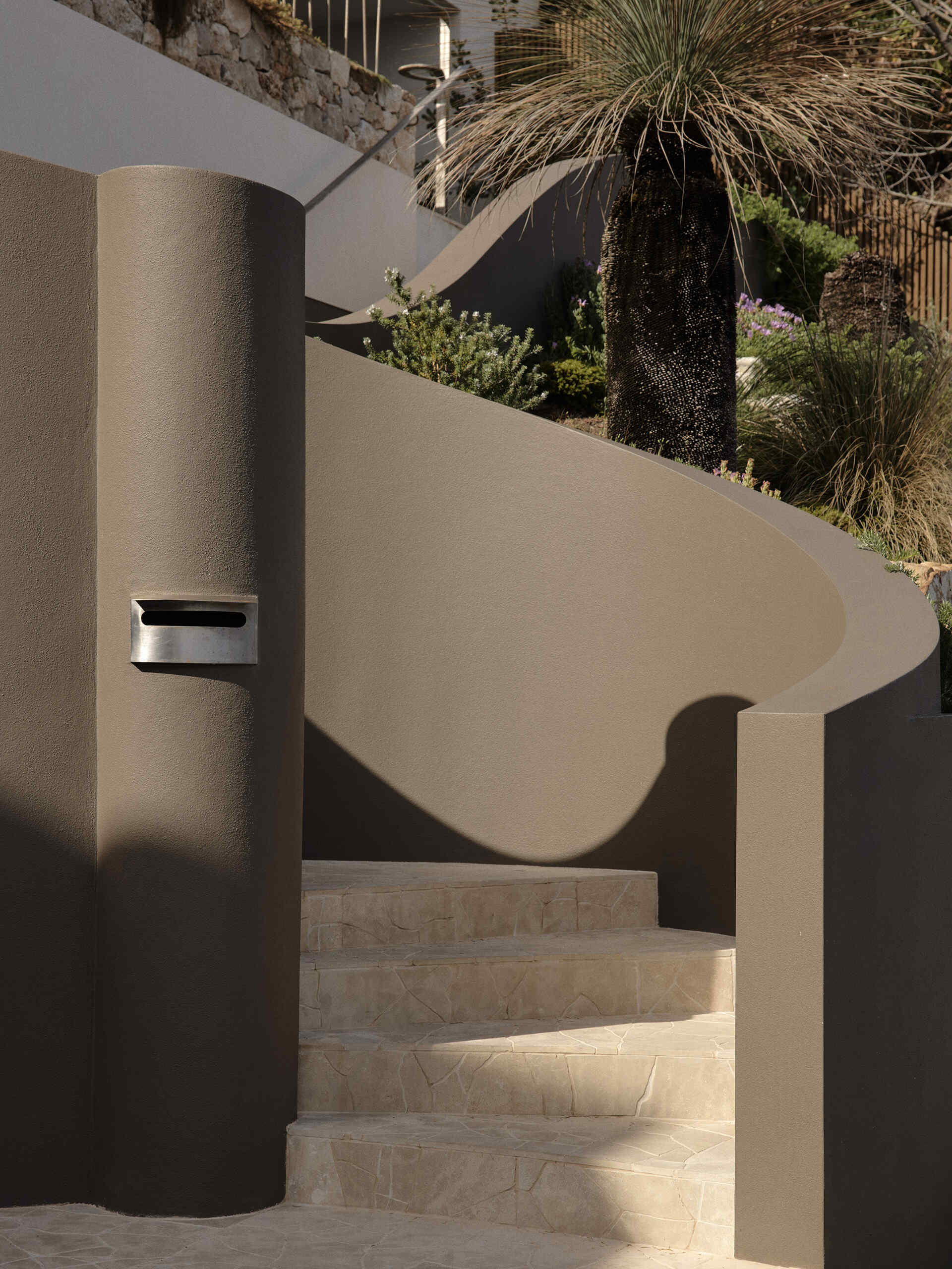 Curved taupe stucco walls, pale stone steps and lush planting create a sculptural entrance to this Byron Bay home, softened by a mature grass tree and touches of purple flowers.