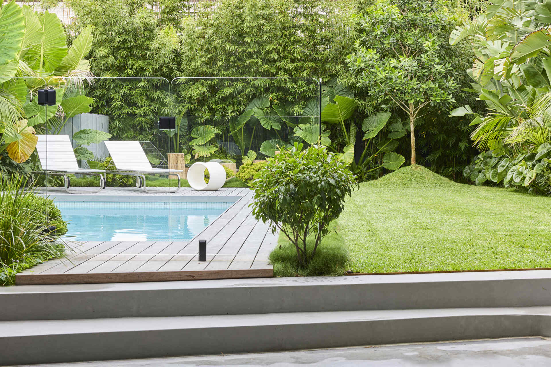A contemporary swimming pool sits within the lush garden at Sasso Nero, framed by timber decking, clear glass screening, white loungers and dense green planting. See how the pool becomes part of the home's outdoor spaces.