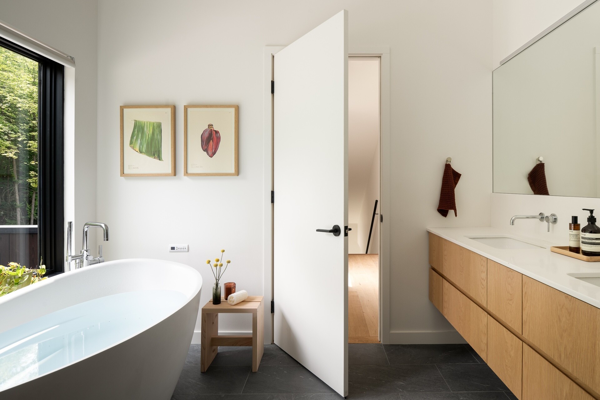 The primary bathroom is bright and pared back, with a freestanding white bathtub positioned beside a large window looking into the trees. White walls and fixtures keep the room feeling fresh and open, while the dark floor adds contrast, and a oak vanity runs along one wall beneath a large mirror.