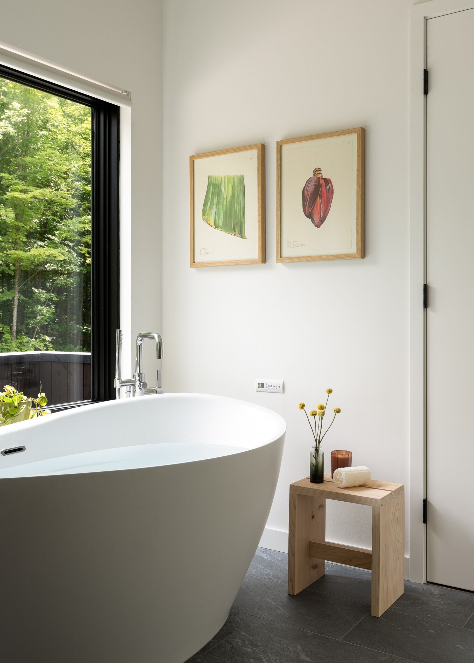 The primary bathroom is bright and pared back, with a freestanding white bathtub positioned beside a large window looking into the trees. White walls and fixtures keep the room feeling fresh and open, while the dark floor adds contrast, and a oak vanity runs along one wall beneath a large mirror.
