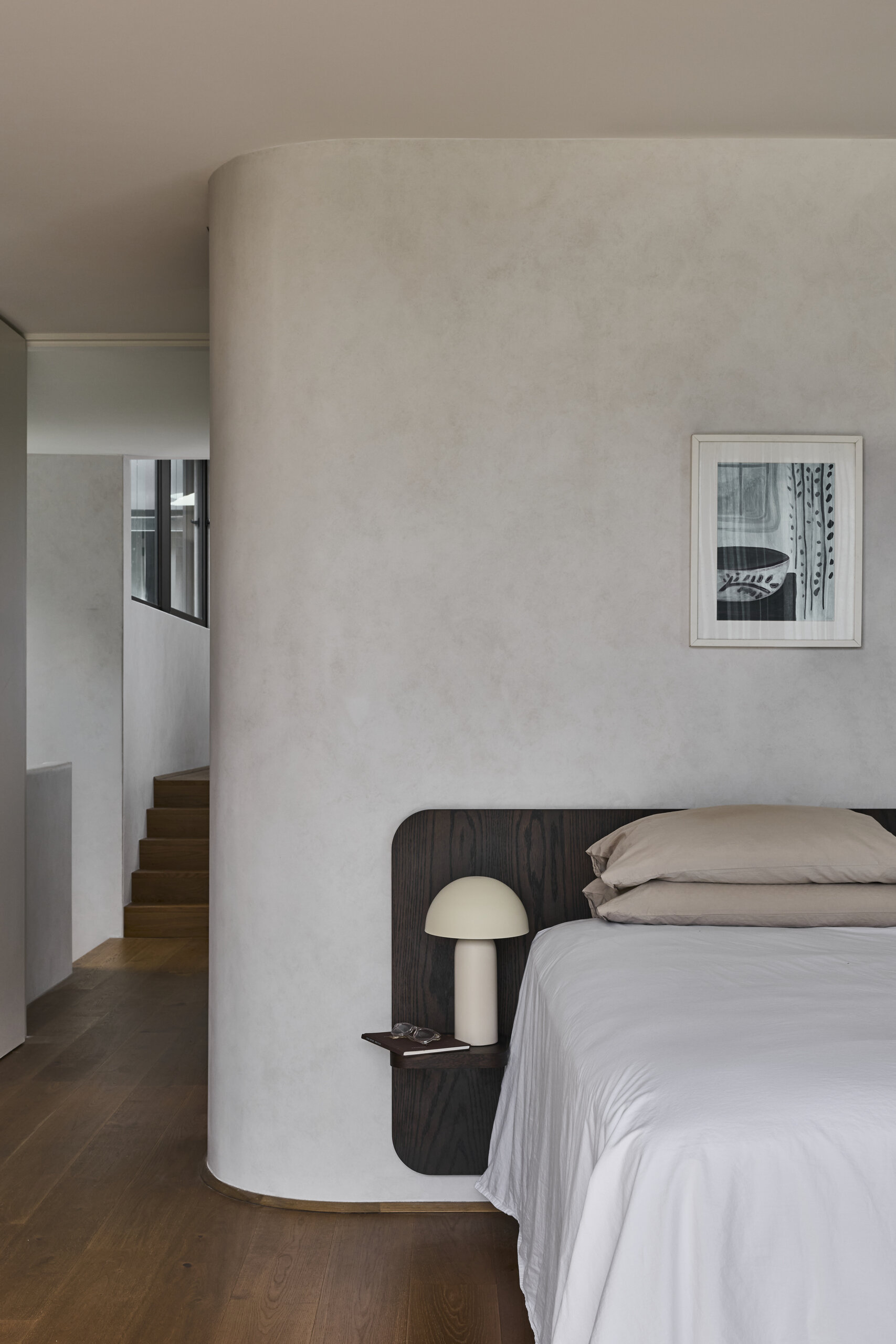 Step inside the primary bedroom at Sasso Nero, featuring a softly curved plaster wall, dark timber headboard, neutral bedding and warm wood floors. The restrained palette creates a calm contemporary interior.