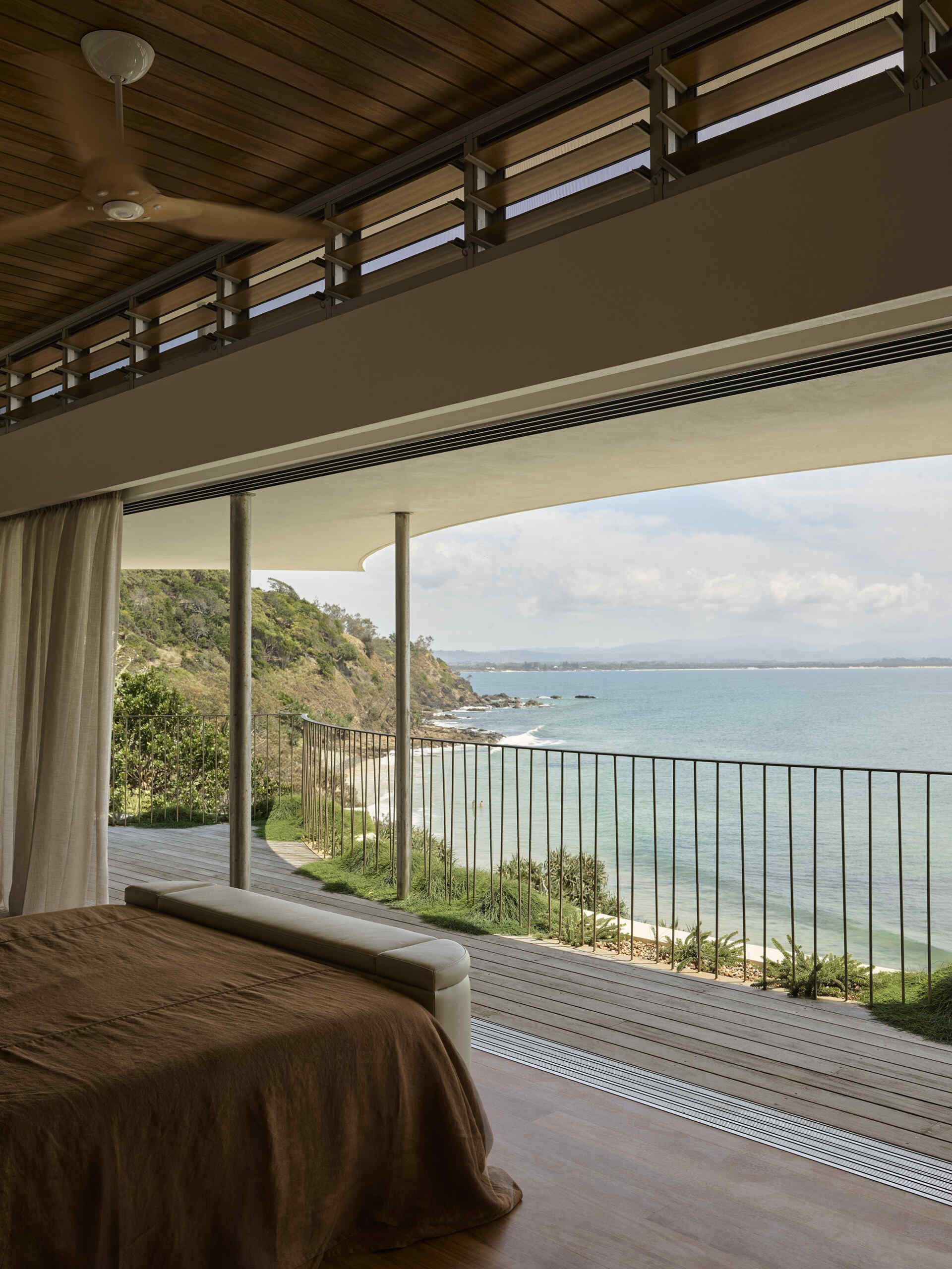 Explore the elevated primary bedroom of this Byron Bay home, featuring warm timber, earthy finishes, mirrored surfaces and large openings that frame the expansive coastal outlook.