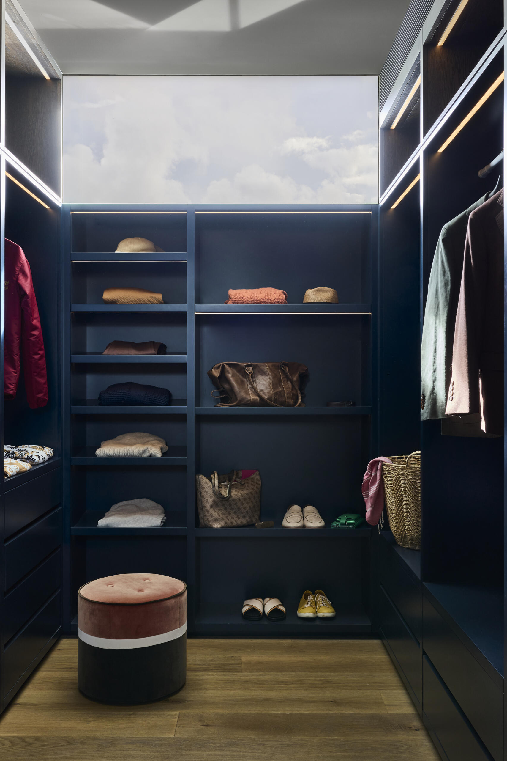 Discover the navy walk-in closet at Sasso Nero, featuring deep blue cabinetry, integrated lighting, warm timber floors, a blush velvet stool and a generous skylight bringing natural light into the space.