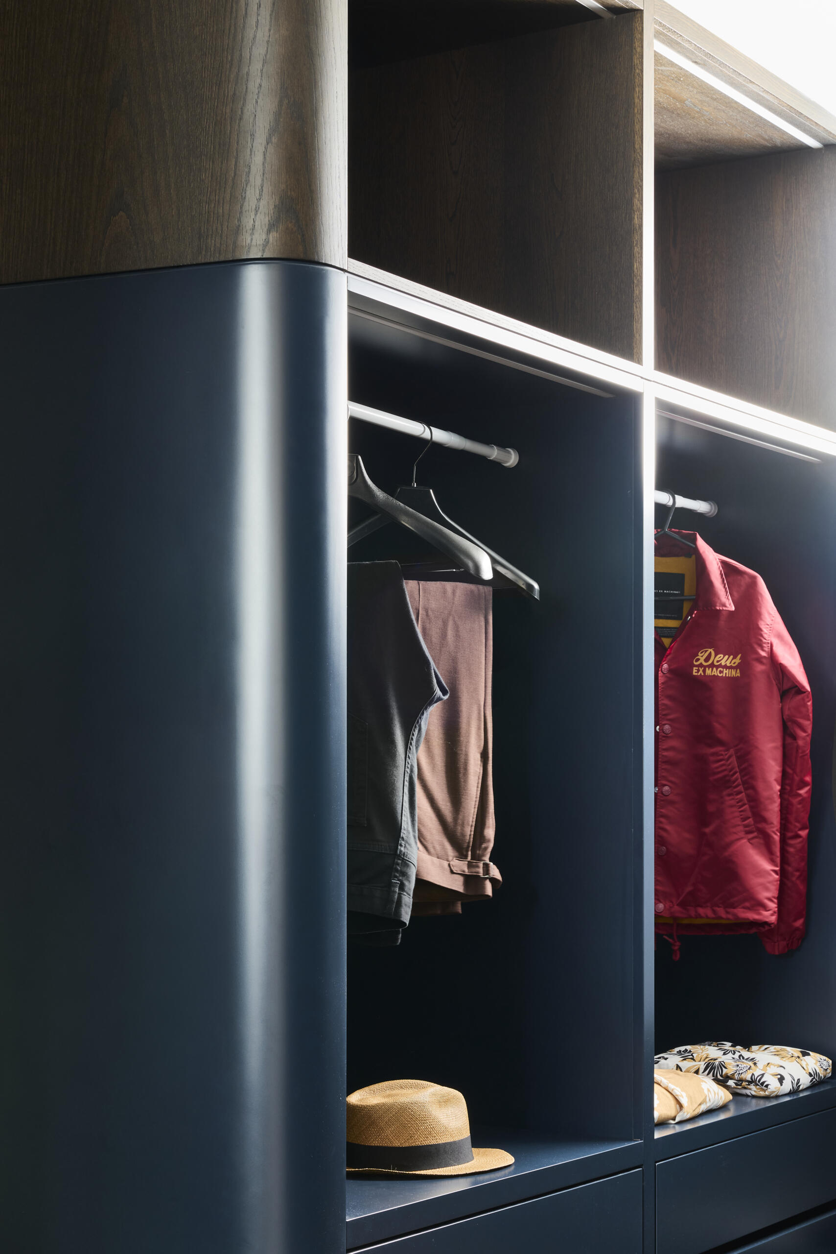 Discover the navy walk-in closet at Sasso Nero, featuring deep blue cabinetry, integrated lighting, warm timber floors, a blush velvet stool and a generous skylight bringing natural light into the space.