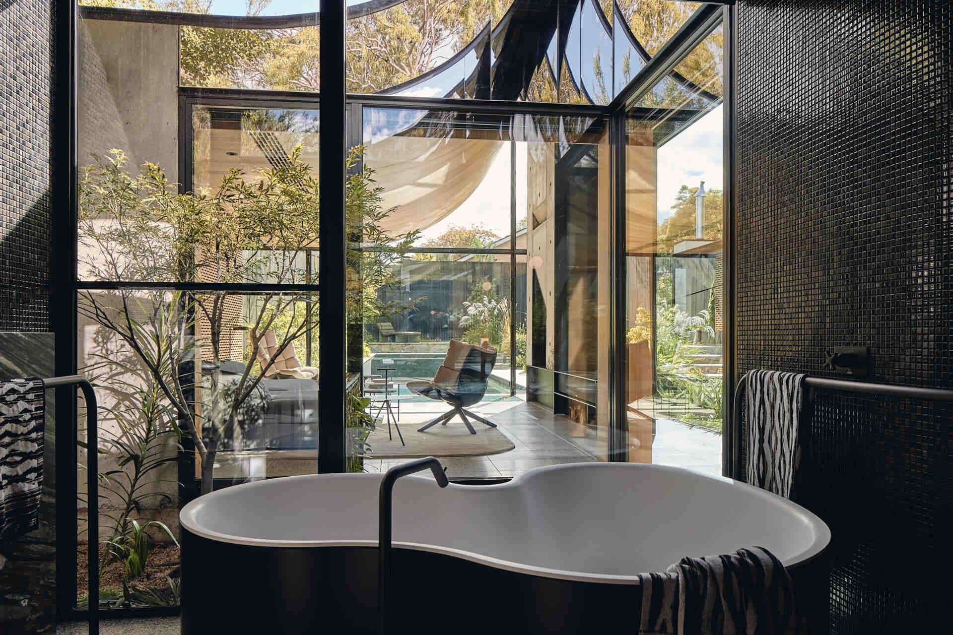 A primary bathroom combines warm wood paneling, dark mosaic tiles and richly veined green stone, with expansive garden views, a sculptural freestanding bath and adjoining dressing area.