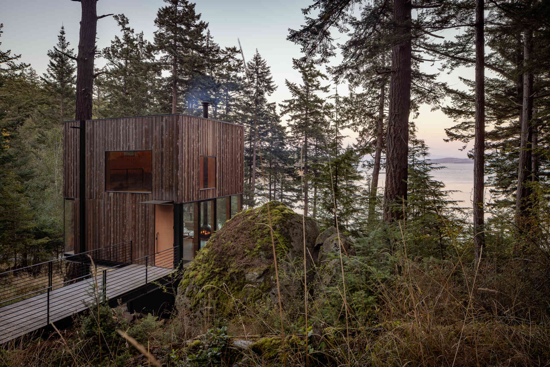 A suspended metal walkway winds through the forest to this tiny Blakely Island cabin, revealing views of Thatcher Bay before reaching the fir entry door.